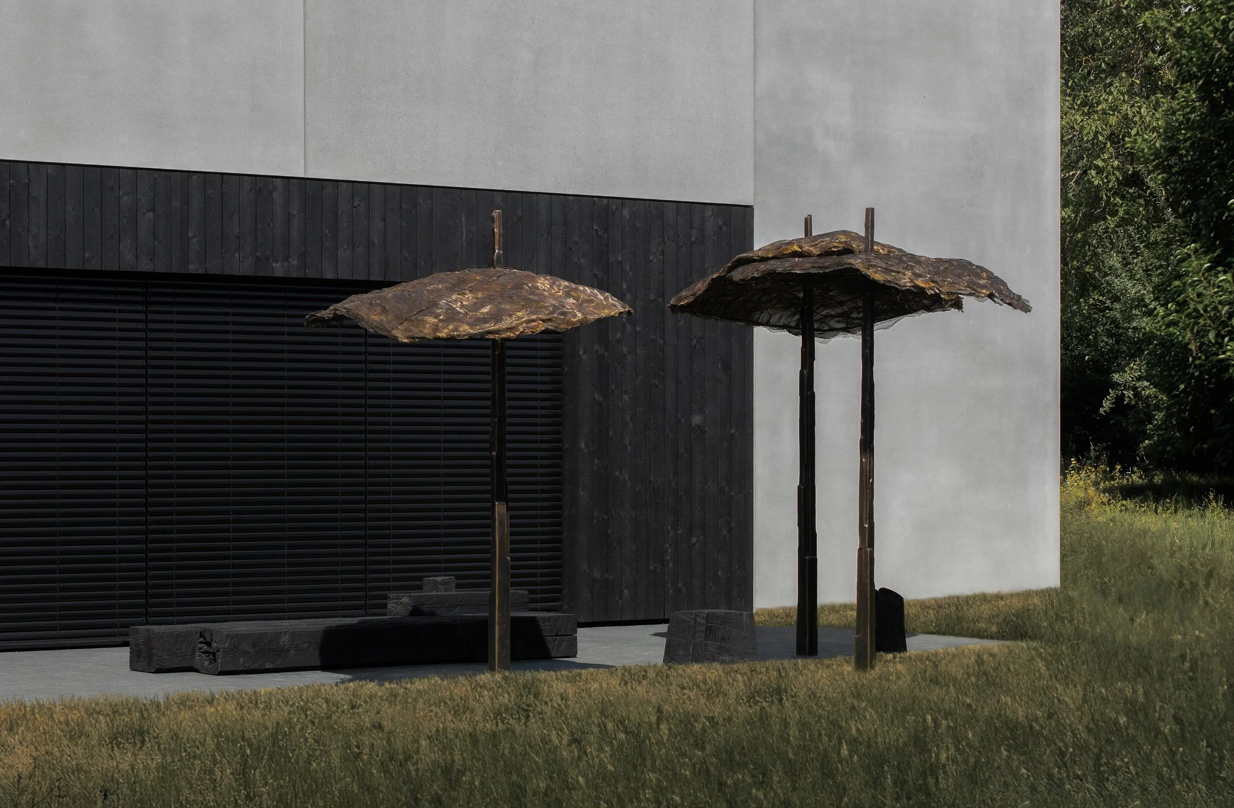 Two outdoor umbrellas made of dark, natural-looking material in front of a modern building with gray walls and black siding.