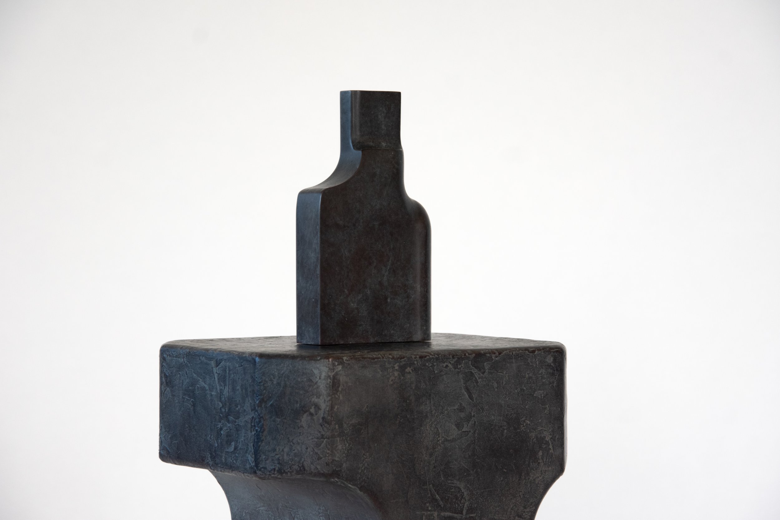 A black sculptural object with a rectangular shape and curved top, placed on a black textured pedestal against a plain white background.