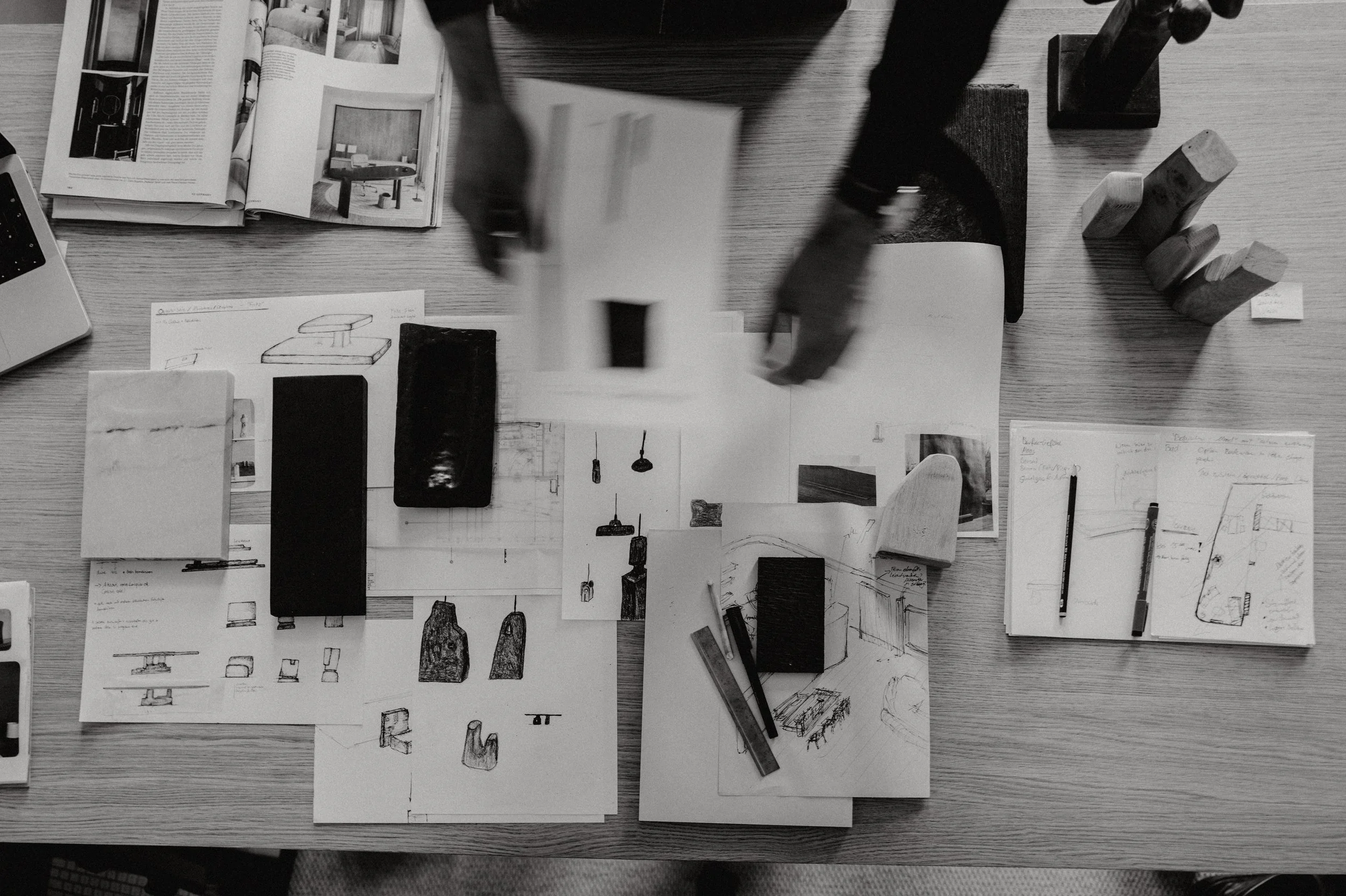 Blueprints, sketches, and design materials for furniture or interior decor spread on a table, with a person’s hands handling a sheet of paper.