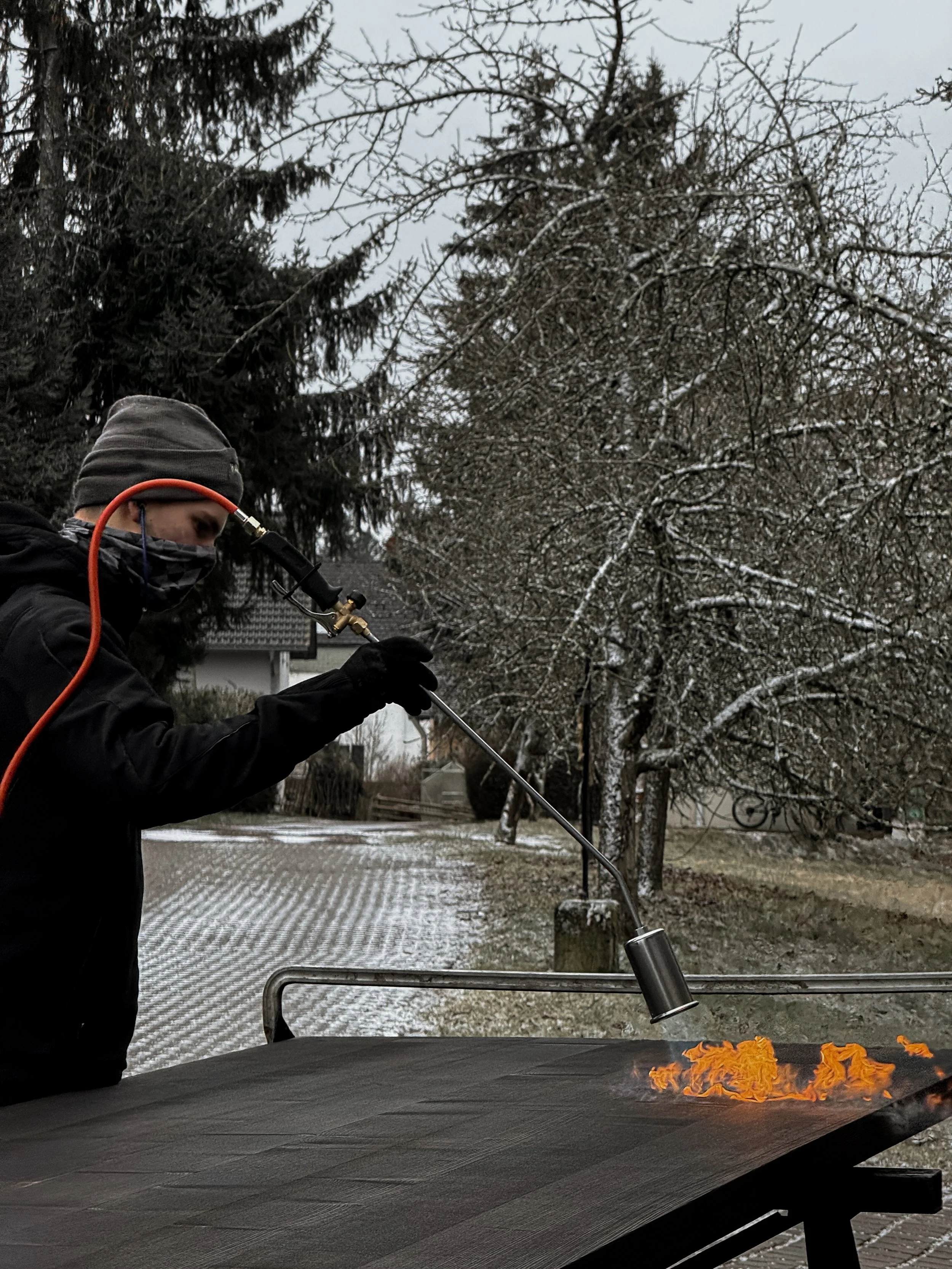 Person in winter clothing and face mask using a blowtorch on a dark surface outdoors on a cold, snowy day.