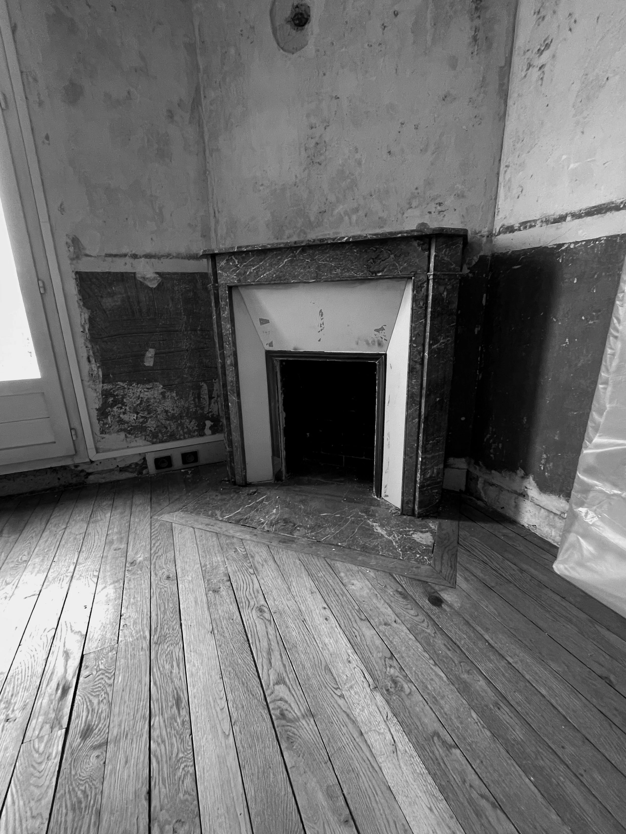 Black and white photo of an empty fireplace with a marble hearth, surrounded by unfinished walls and a wooden floor with visible grain and gaps between the planks.