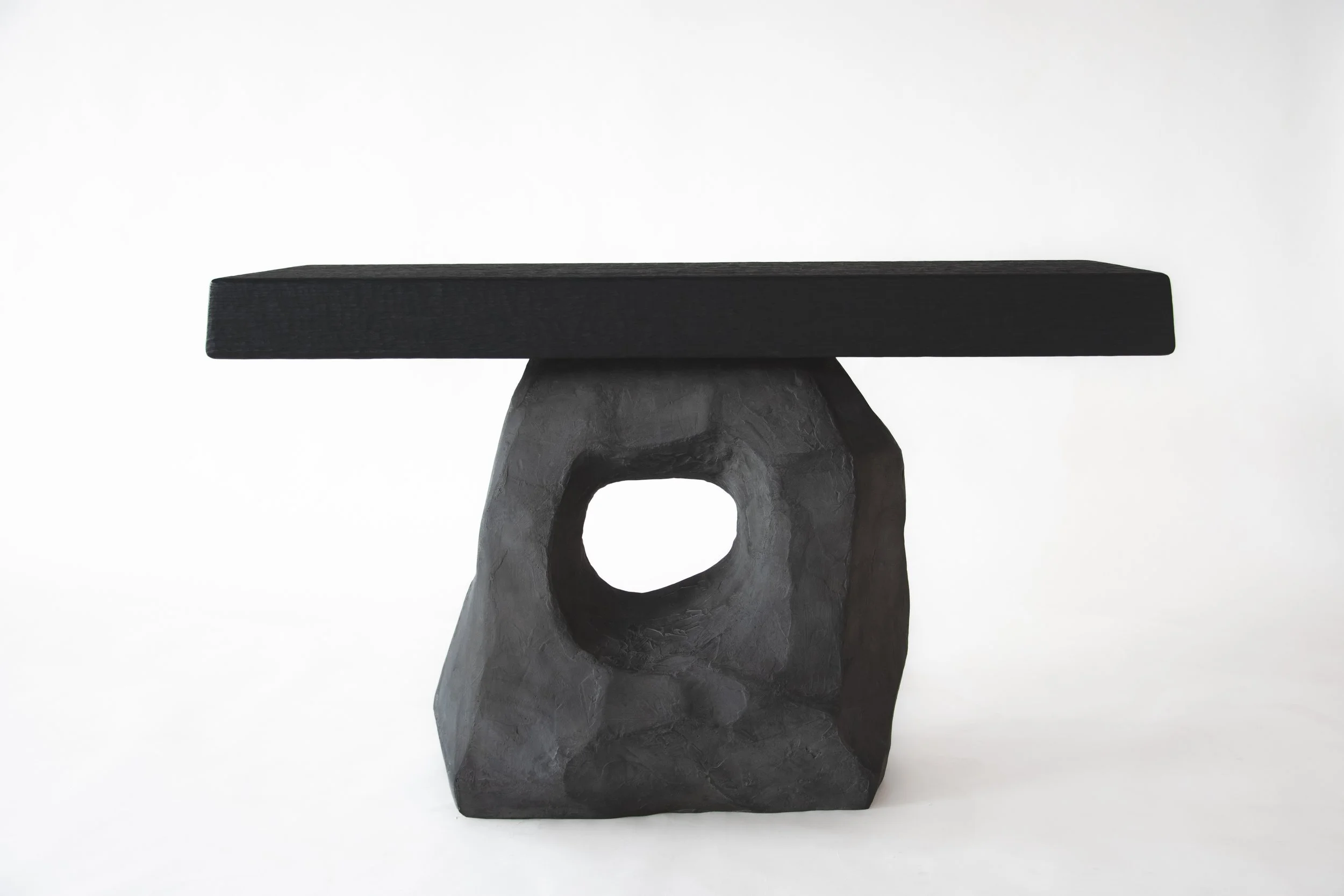 A black rectangular tabletop on a sculptural black base with a circular hole in the center, set against a white background.