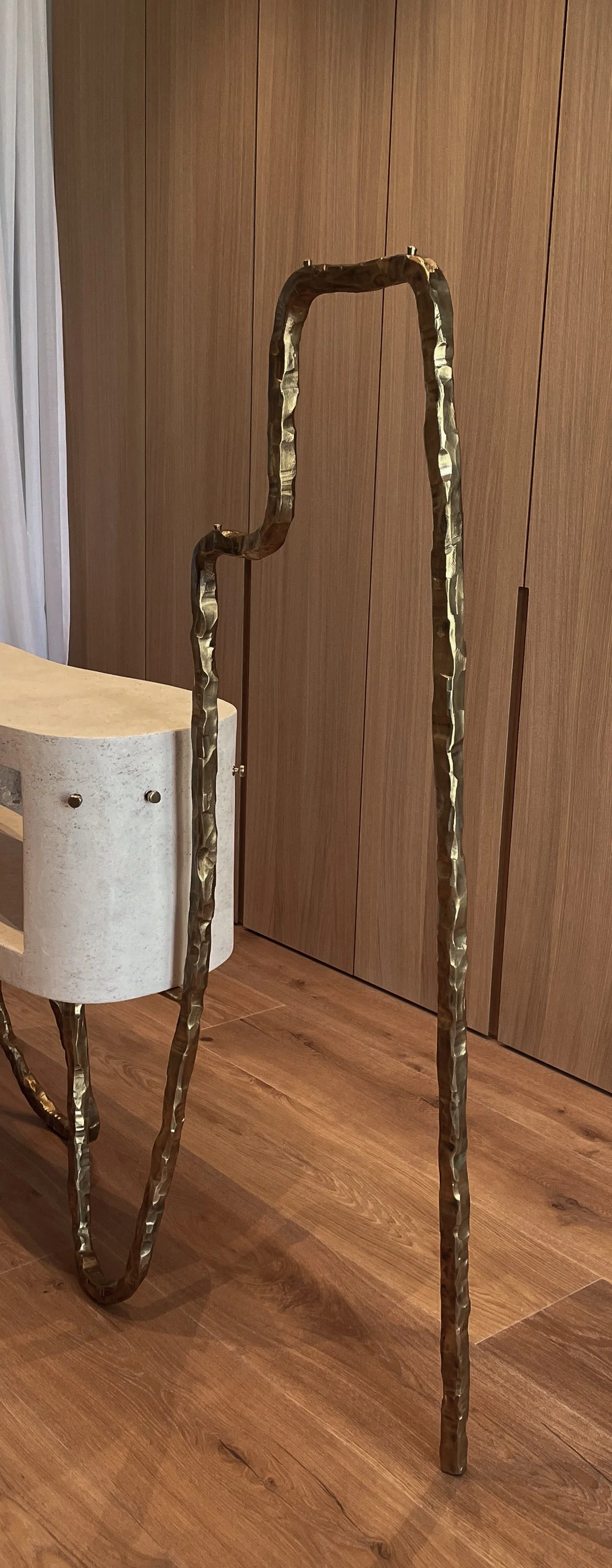 A walking cane with a hammered metallic finish, standing upright near a wooden closet door and a curved white concrete table.