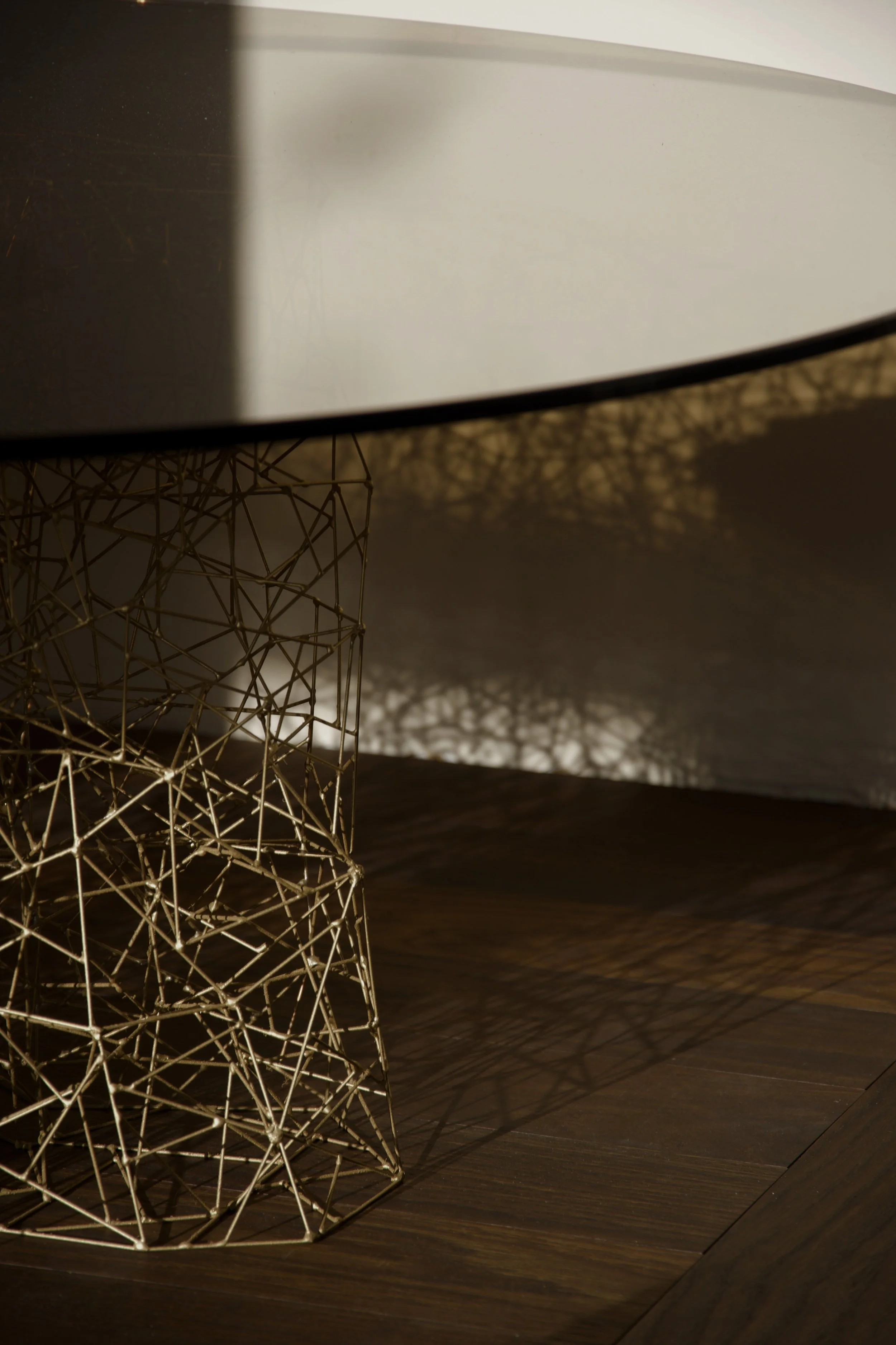 Close-up of a modern silver metallic table lamp with an intricate geometric wire base, casting shadows on the wooden surface beneath, with a white lampshade partially visible.