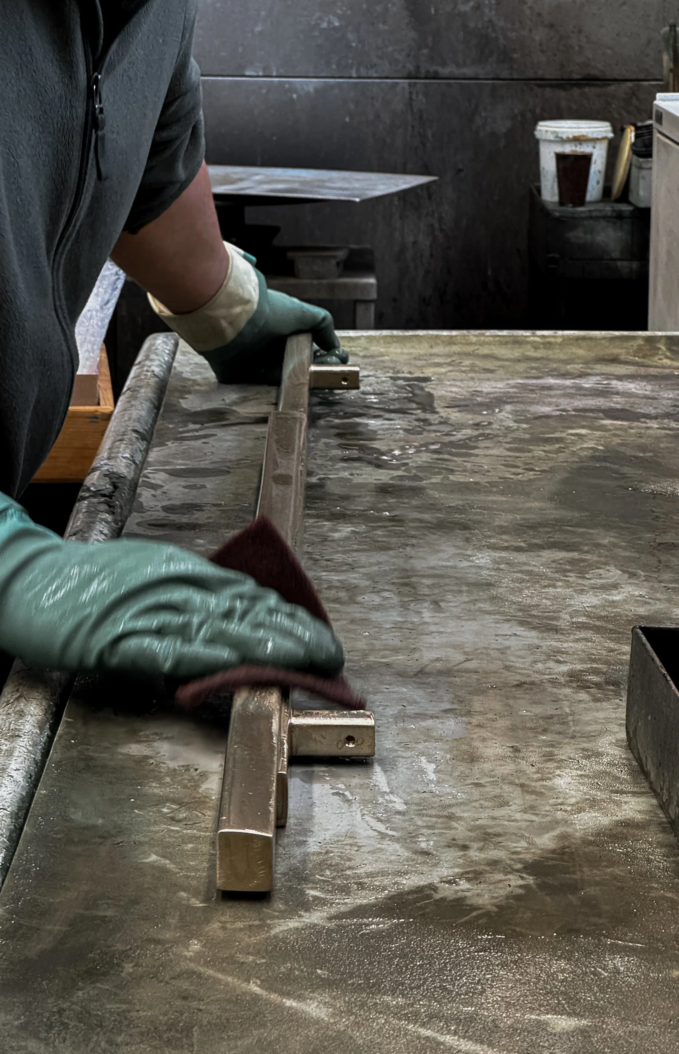 A person wearing green gloves using a sanding tool on a metal surface in a workshop.