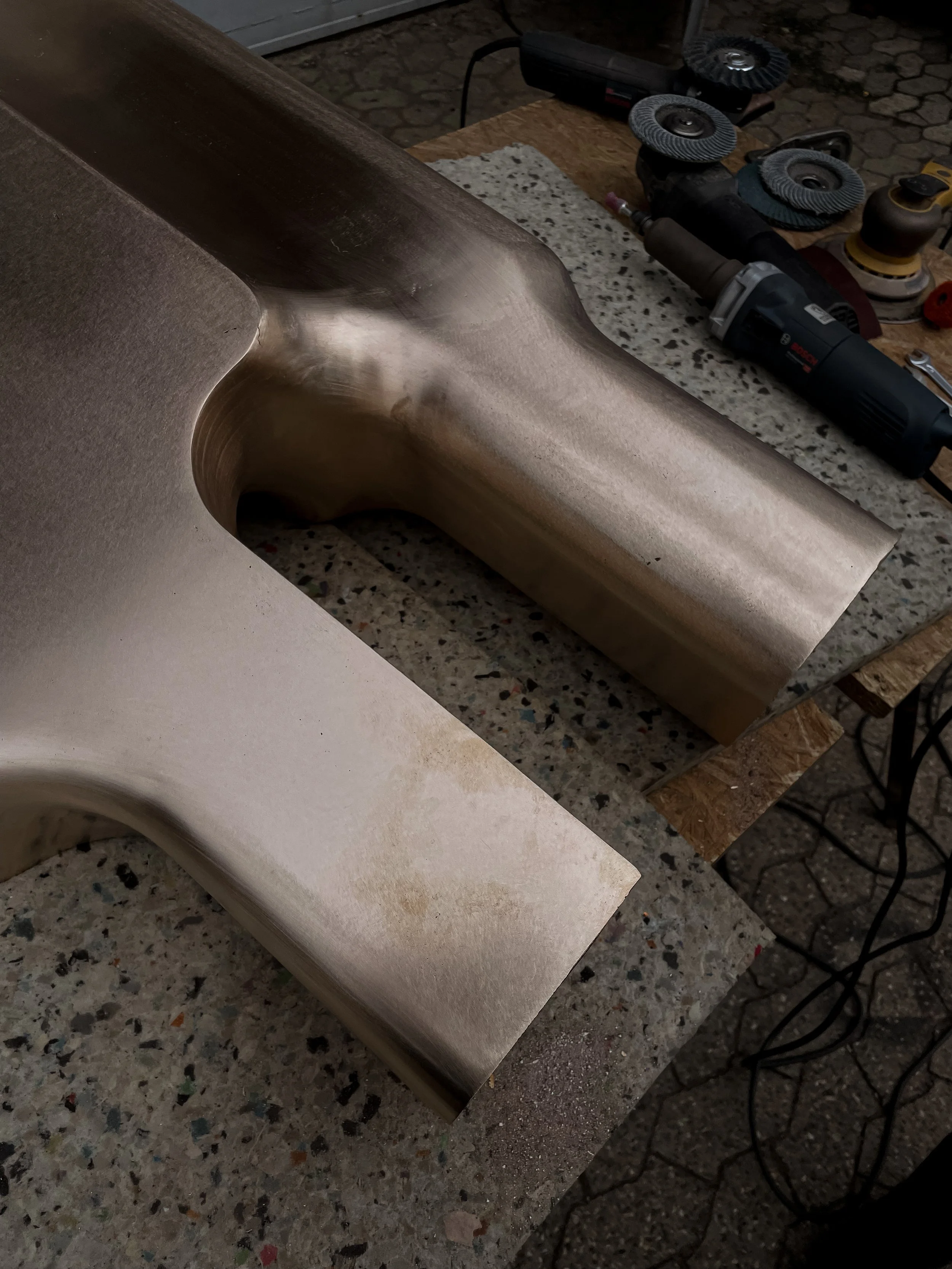 Close-up of two metallic, curved, polished pieces on a workbench, with power tools and grinding discs in the background.