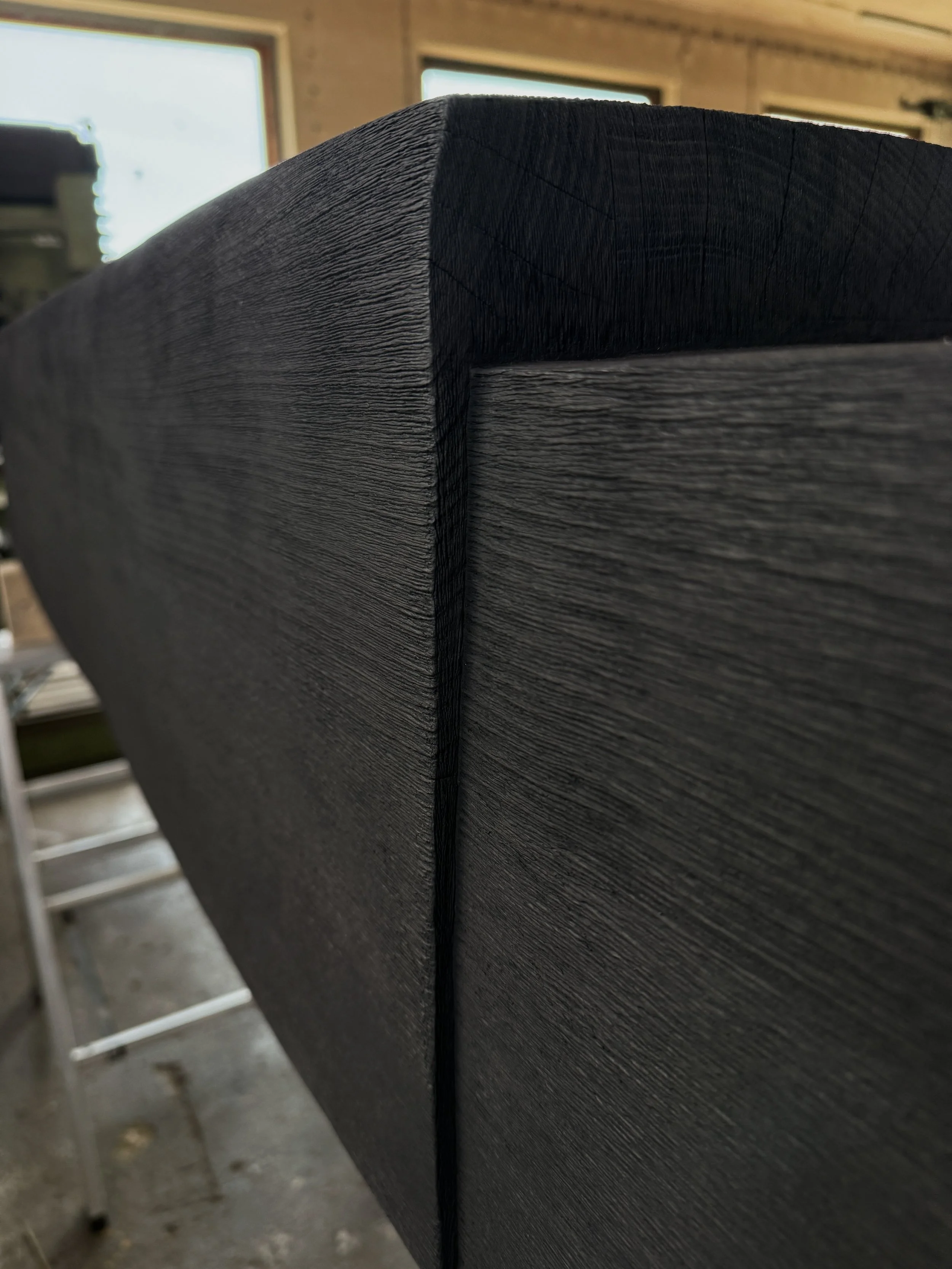 Close-up of a black wooden piece with visible wood grain, indoors with windows in the background.