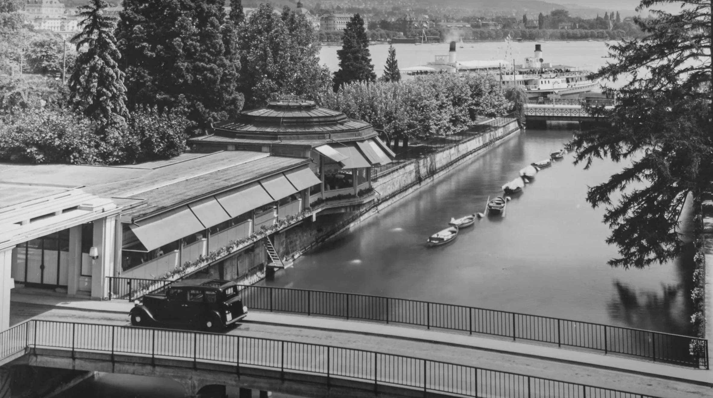 A black and white photo of a riverside scene with boats, trees, and buildings in the background, with a vintage car parked on a bridge.