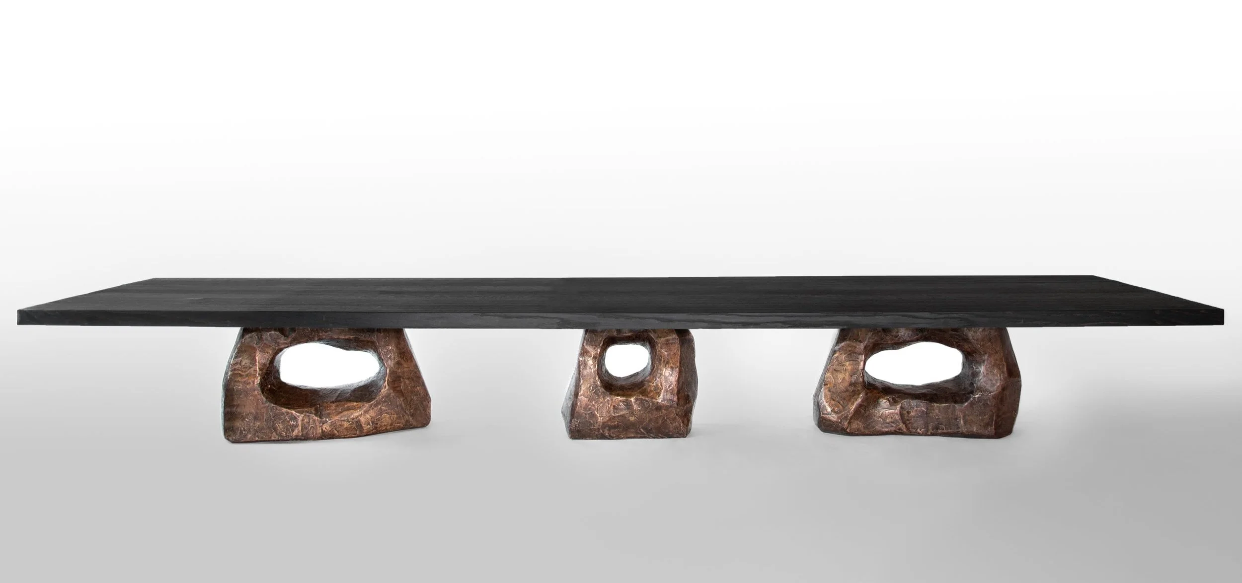 Long black wooden table supported by three irregularly shaped bronze legs with hollow centers, set against a plain white background.
