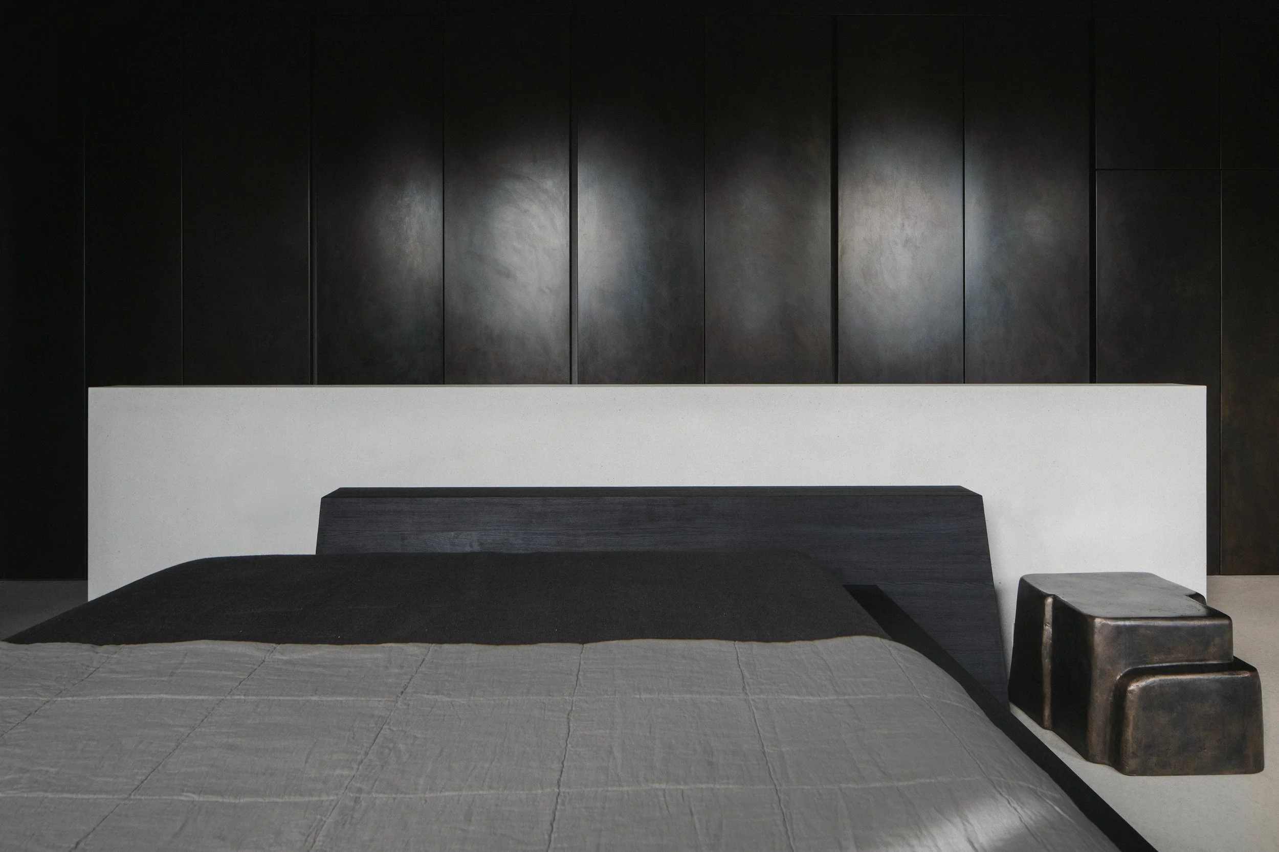 Minimalist bedroom with a bed featuring a dark headboard, a white wall behind the bed, and two metallic stools beside it, with dark paneling on the wall.