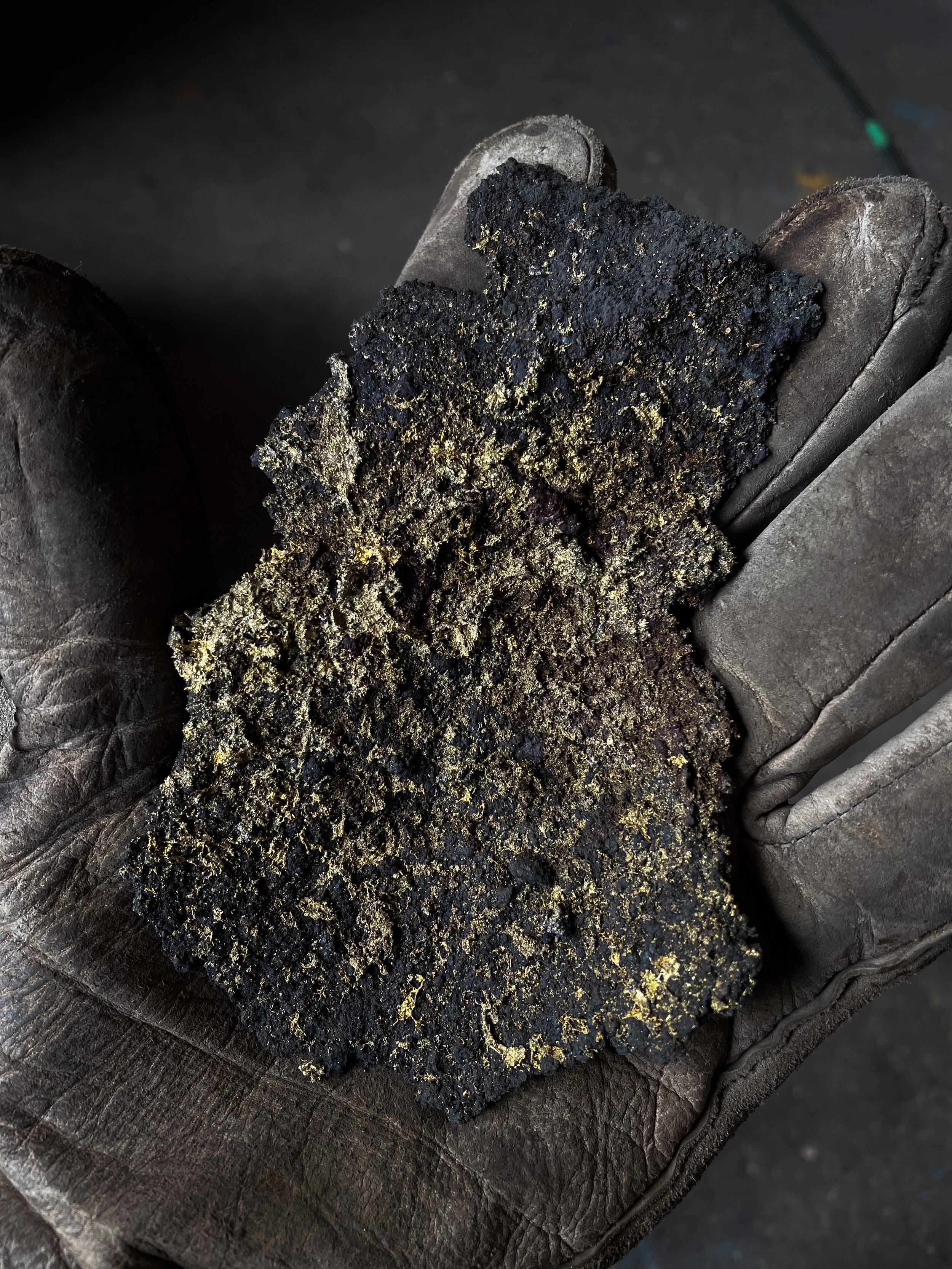 A worker's gloved hand holding a large chunk of molten metal with a rough, black, and gold-textured surface.