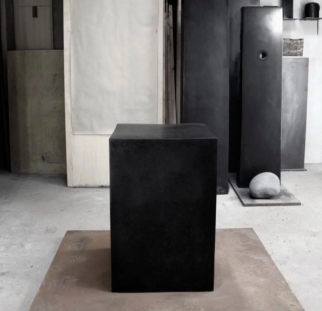 A black rectangular sculpture on a wooden base in an art studio or gallery, with other minimalist sculptures and artworks in the background.