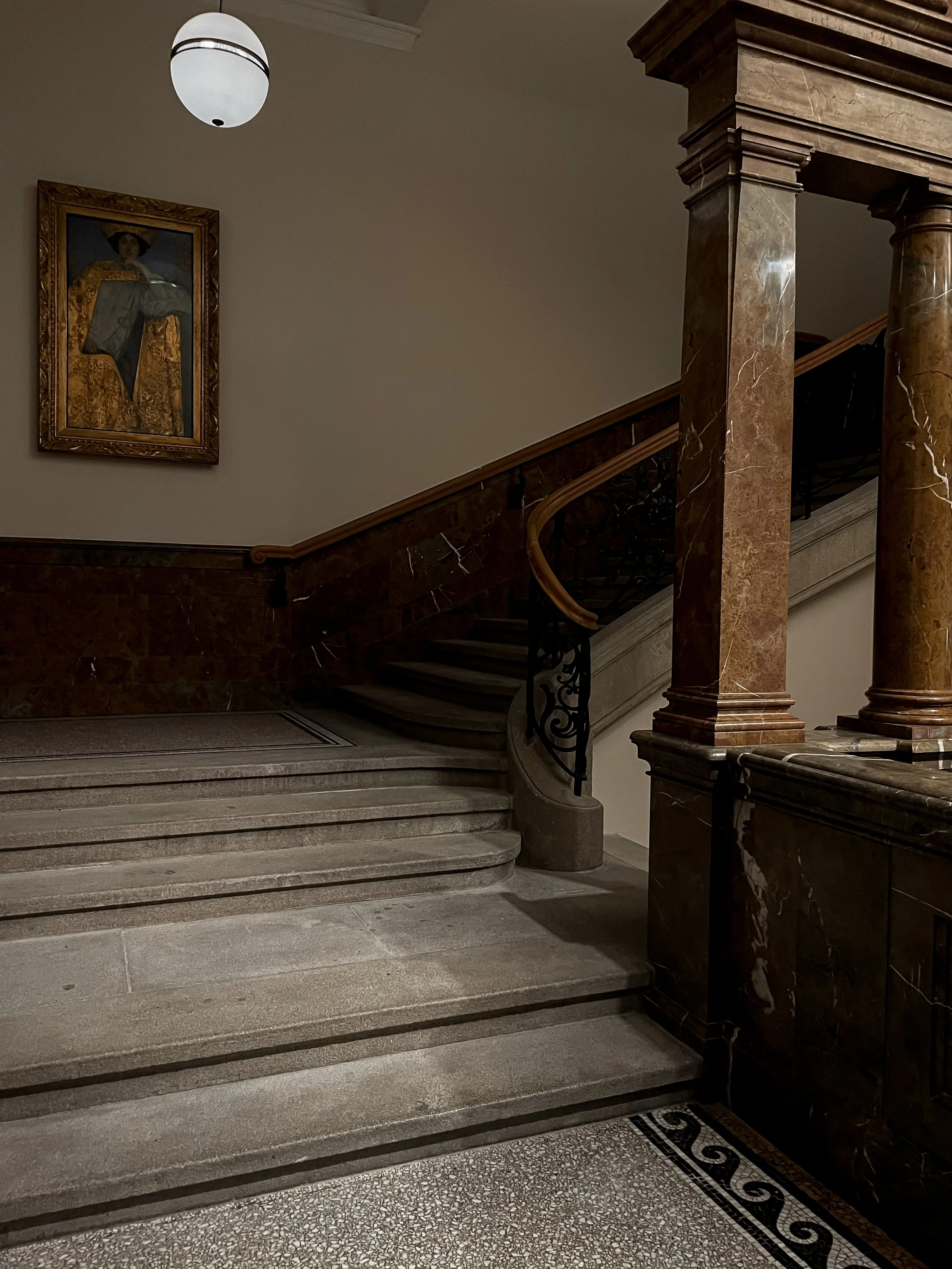 Interior view of a marble staircase with a wooden handrail, black wrought iron balusters, a painting of a woman in a gold frame on a beige wall, and a ceiling light fixture.