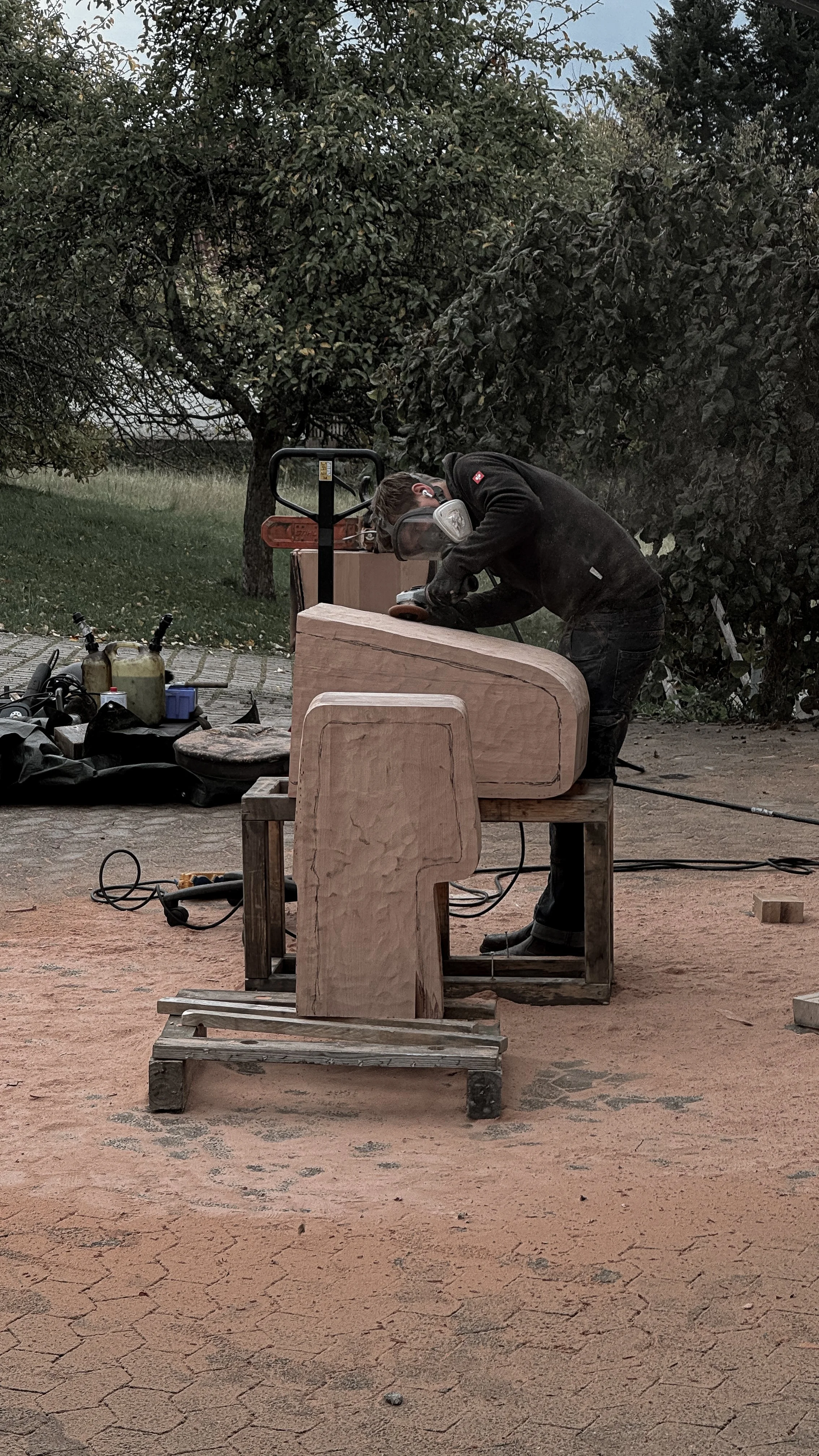 Person woodworking outdoors, cutting a large piece of wood on a workbench with tools and equipment nearby.