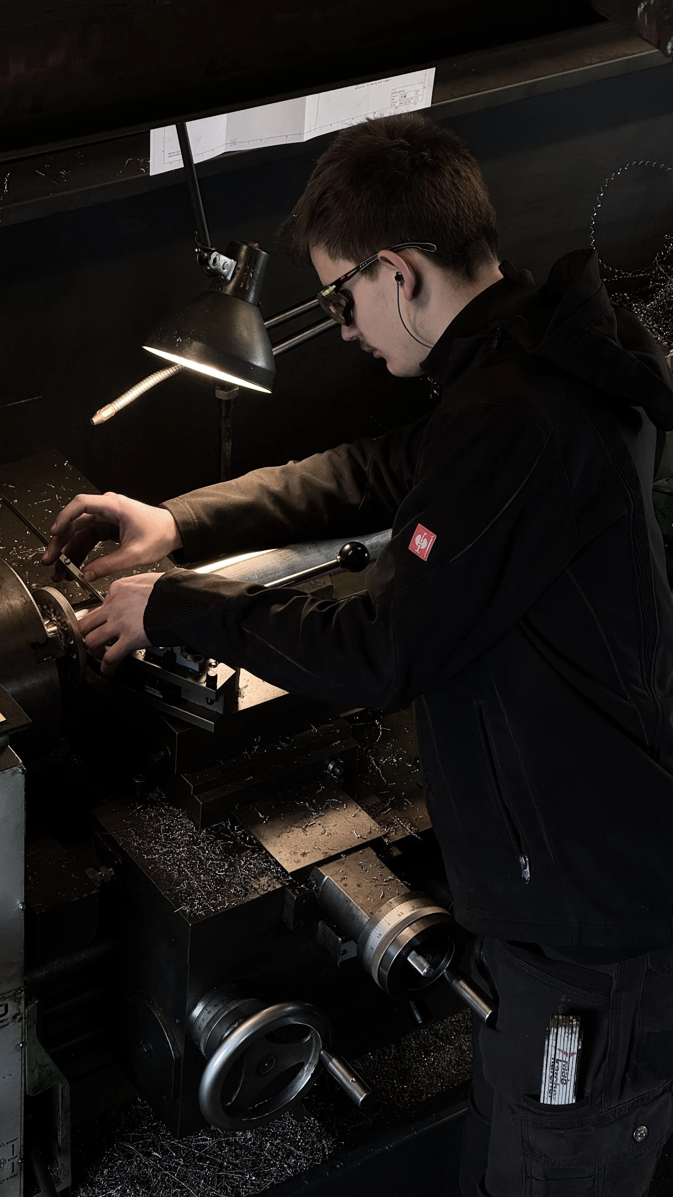 A young man working on a metal lathe machine, wearing safety glasses and earplugs, in a workshop