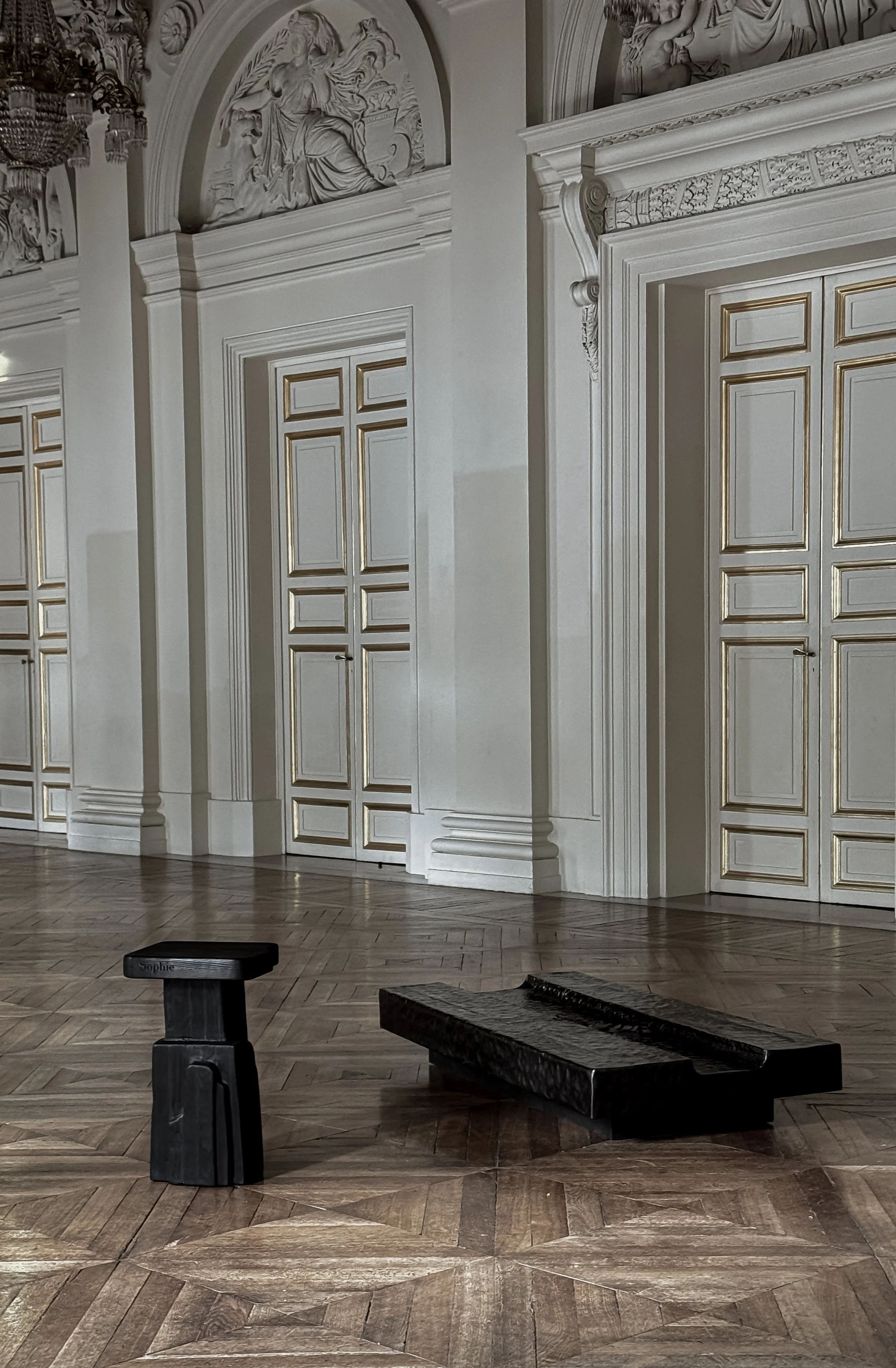 Elegant interior room with ornate white and gold-paneled doors, detailed moldings, and carvings on the walls and ceiling. Two black sculptural benches are on a polished parquet wood floor.