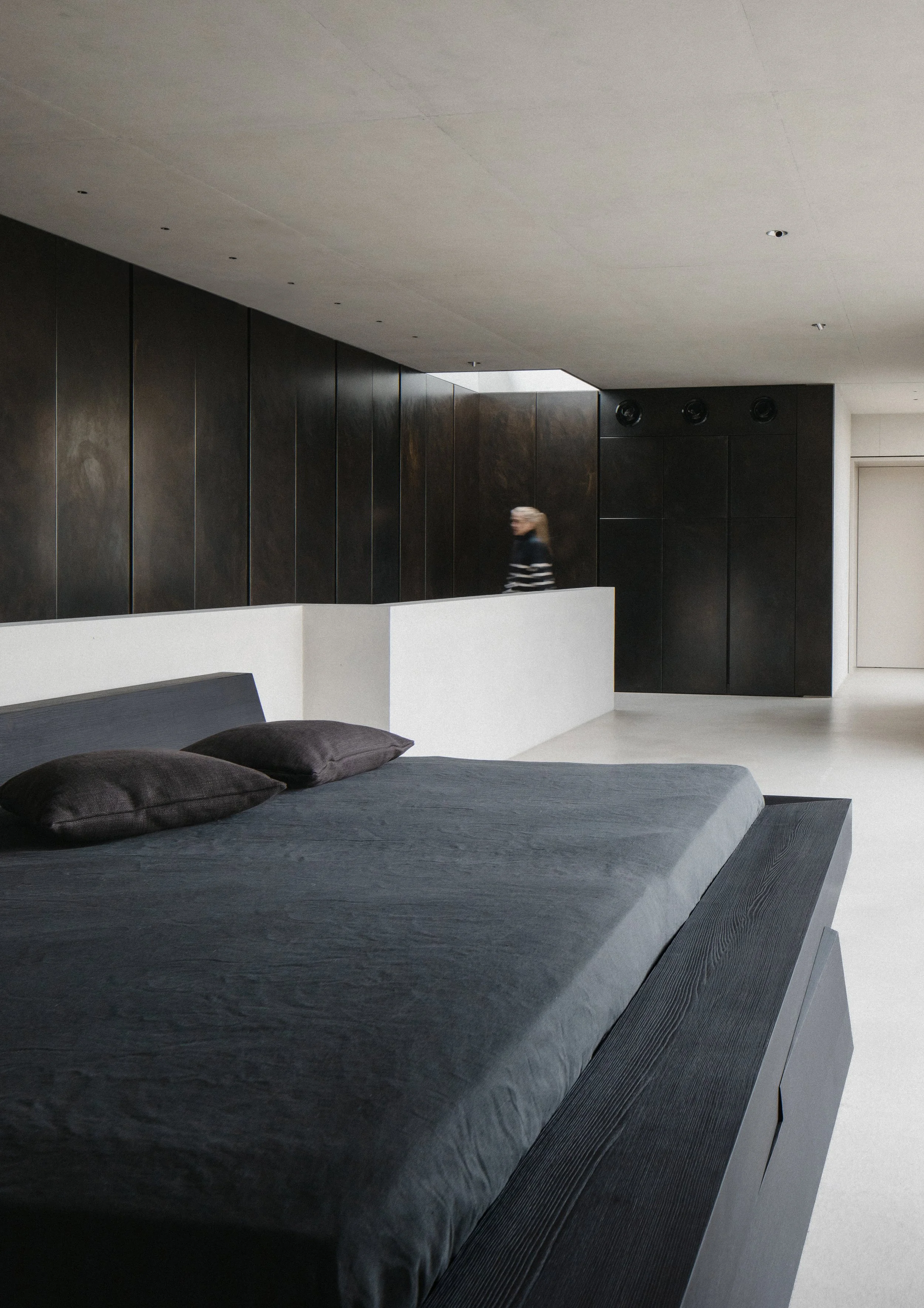 Modern bedroom with dark-colored bed and black pillows, minimalist interior with dark wooden wall panels, white floor, and a woman walking in the background.