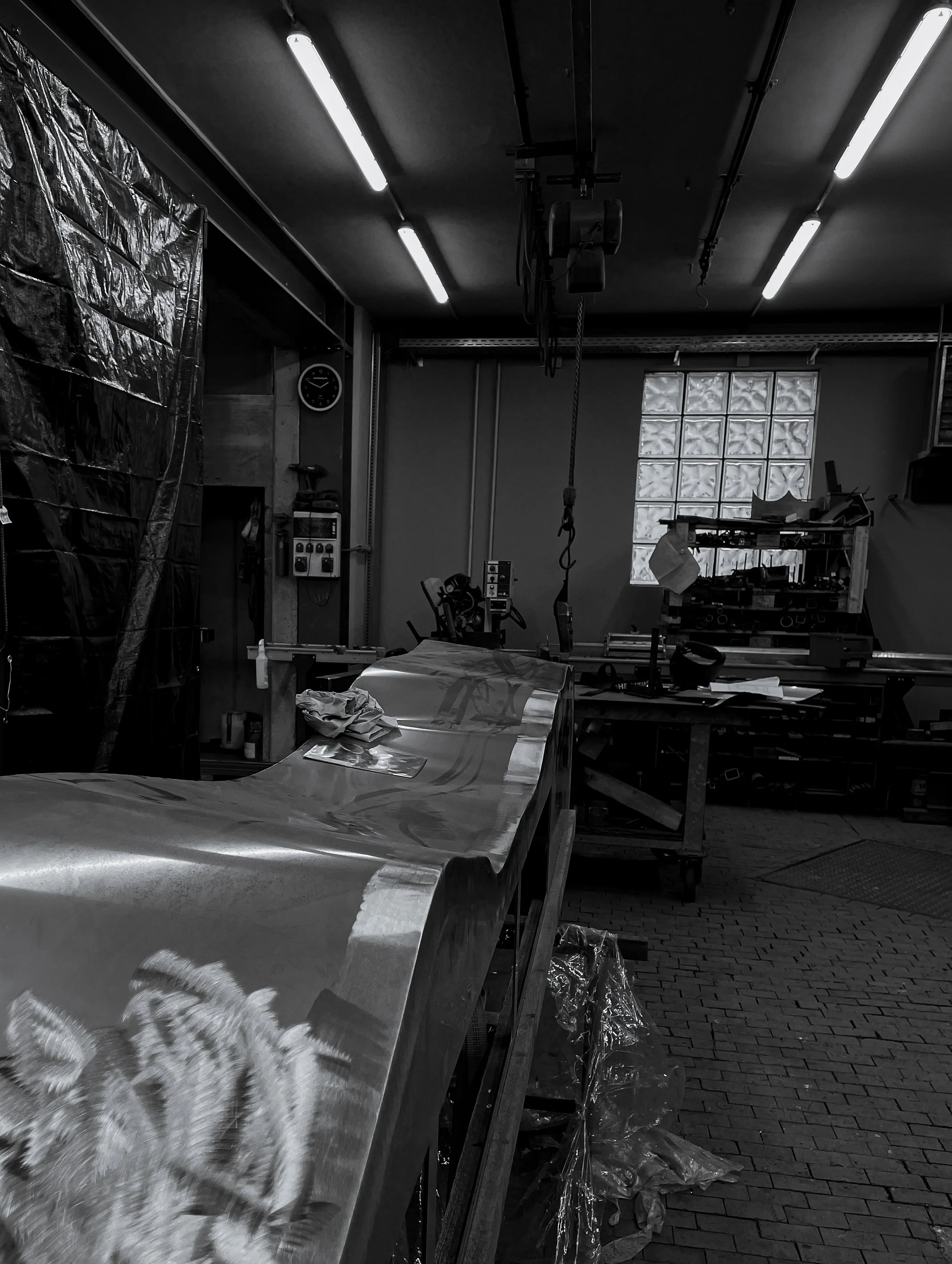A black and white photo of a workshop or fabrication space with a metallic work table, various tools, equipment, and supplies, illuminated by overhead fluorescent lights, with a glass block window in the background.