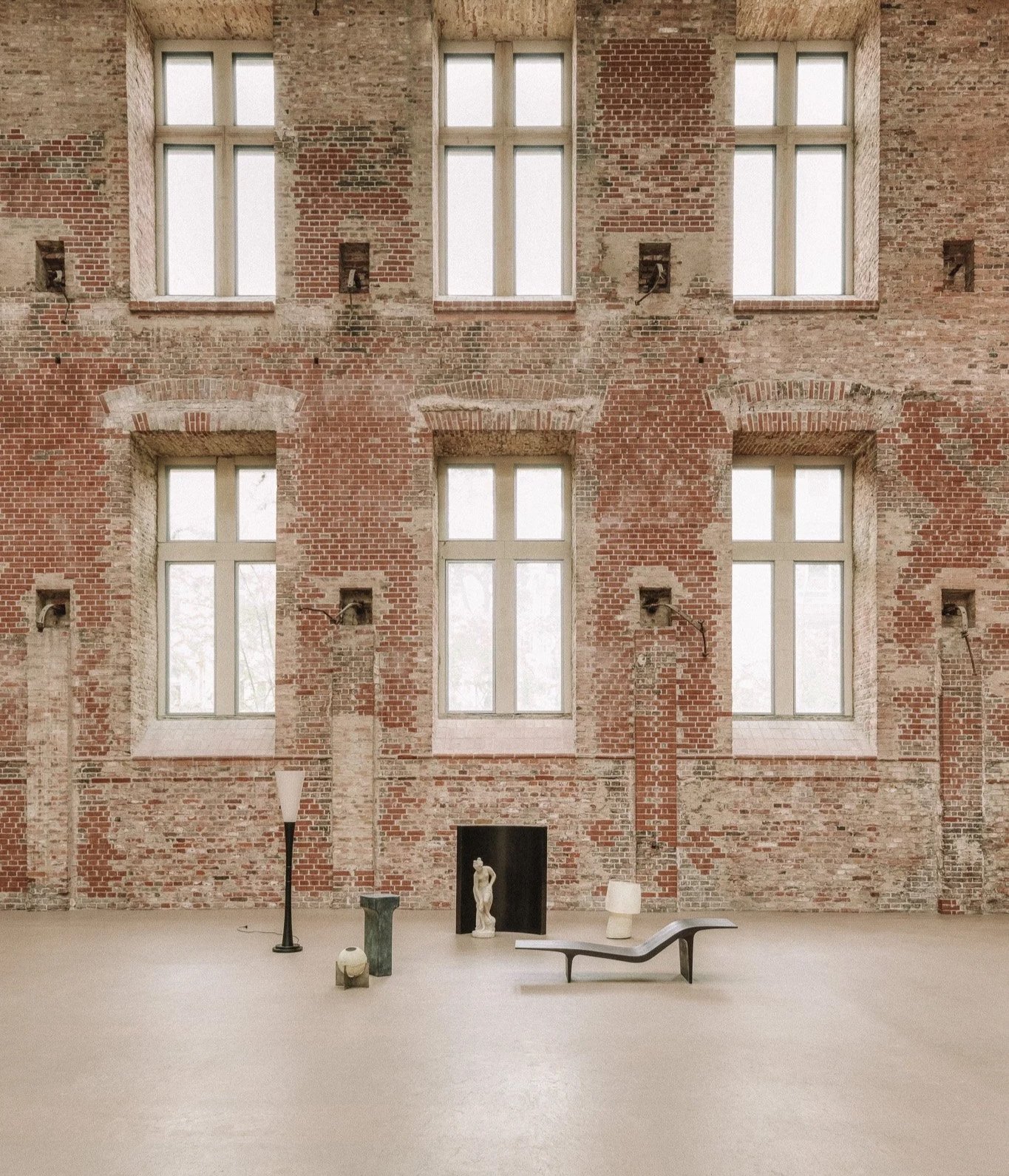 Empty art gallery with exposed brick wall, large windows, and minimal furniture, including a sculpture, a bench, and lamps.