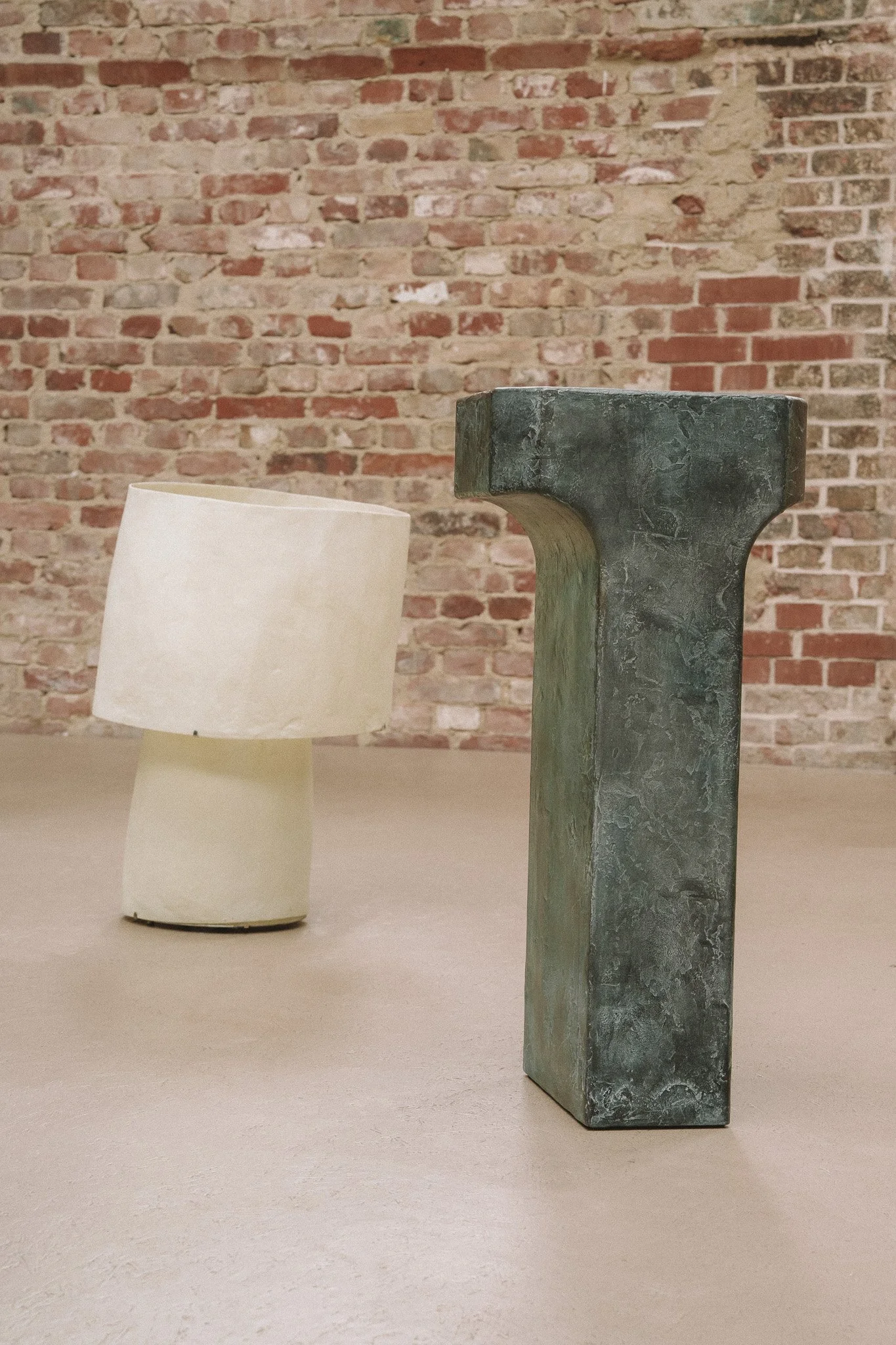 A white, irregularly shaped lamp with a cylindrical base and tilted shade next to a large, dark gray, T-shaped sculpture with a textured surface, both placed on a smooth floor against an exposed brick wall.