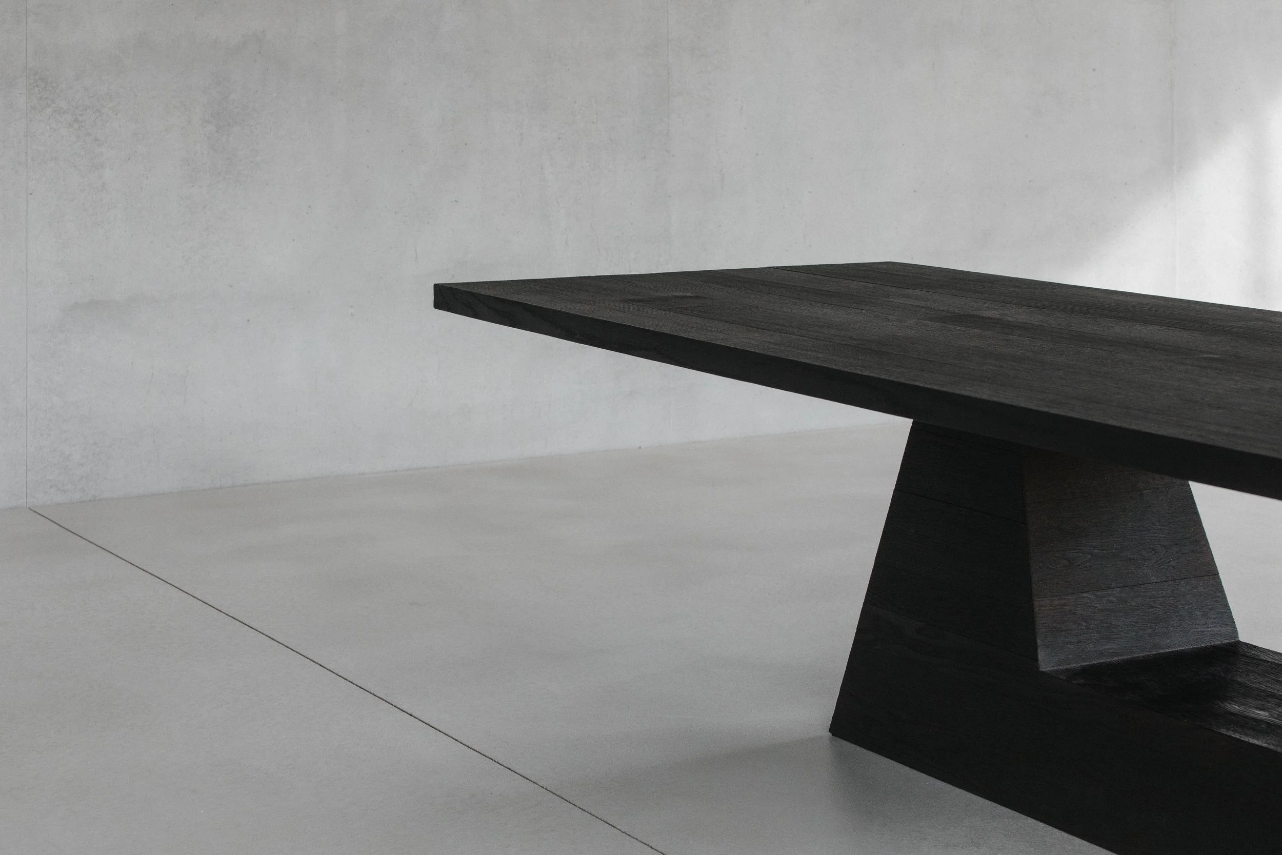 A modern, minimalist black wooden table with a geometric, angled base, set against a plain light gray background.
