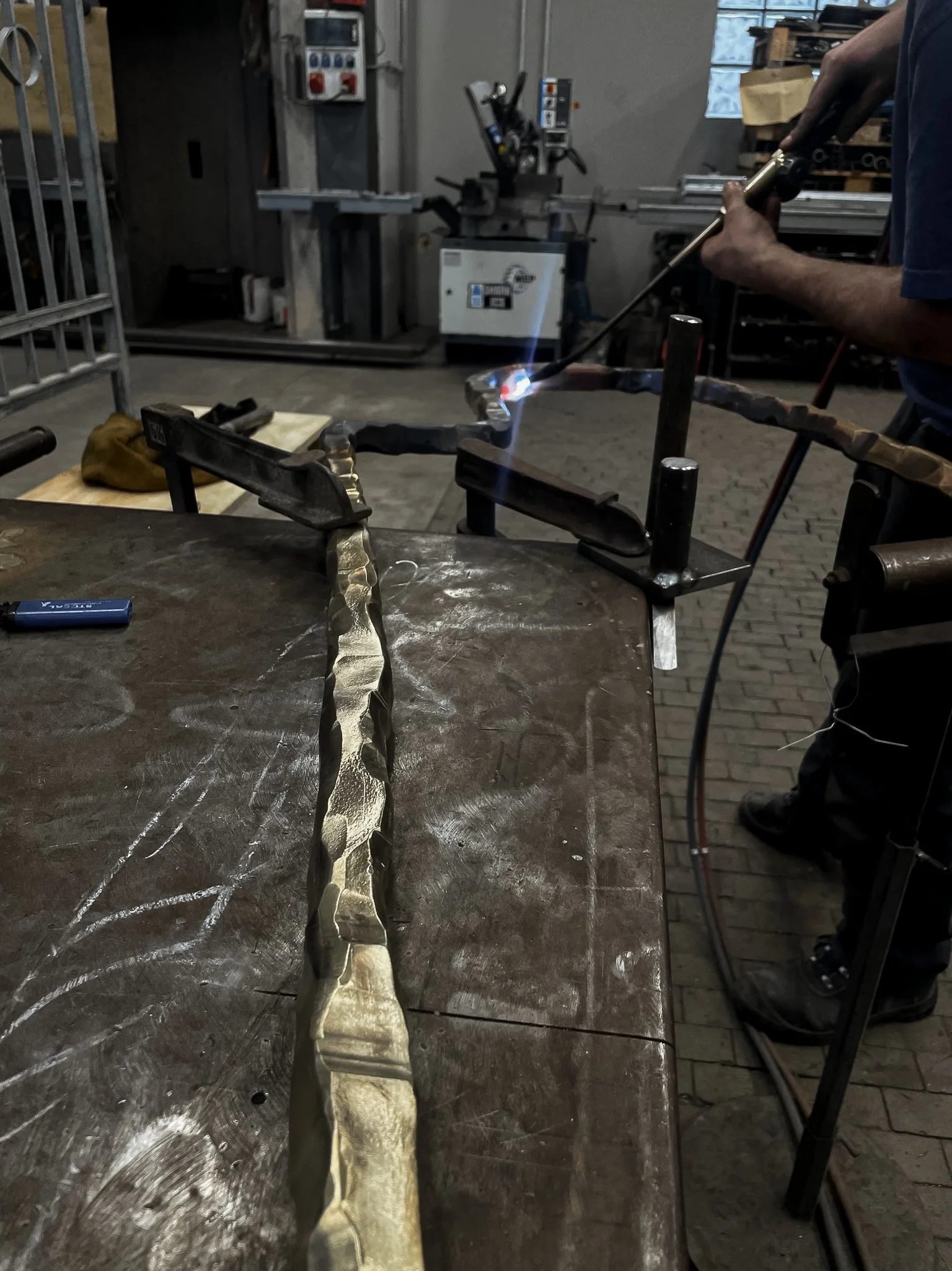 A person welding metallic rods on a work table in a workshop. The welder is using a welding torch, and sparks and bright light are visible at the welding site. Various tools and machinery are in the background.