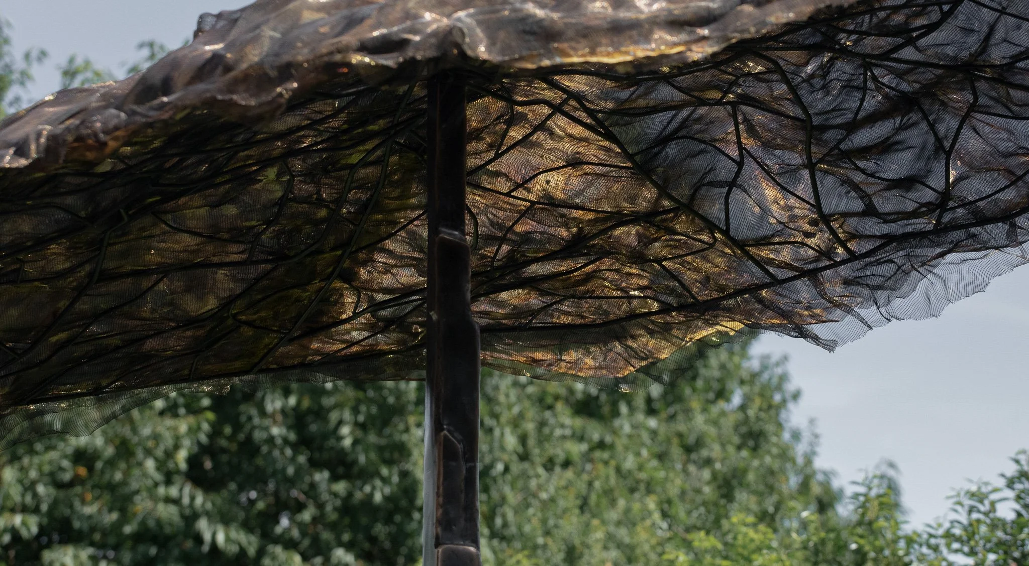 Close-up of a black metal patio umbrella with a brown fabric canopy, set against a background of green trees and a cloudy sky.