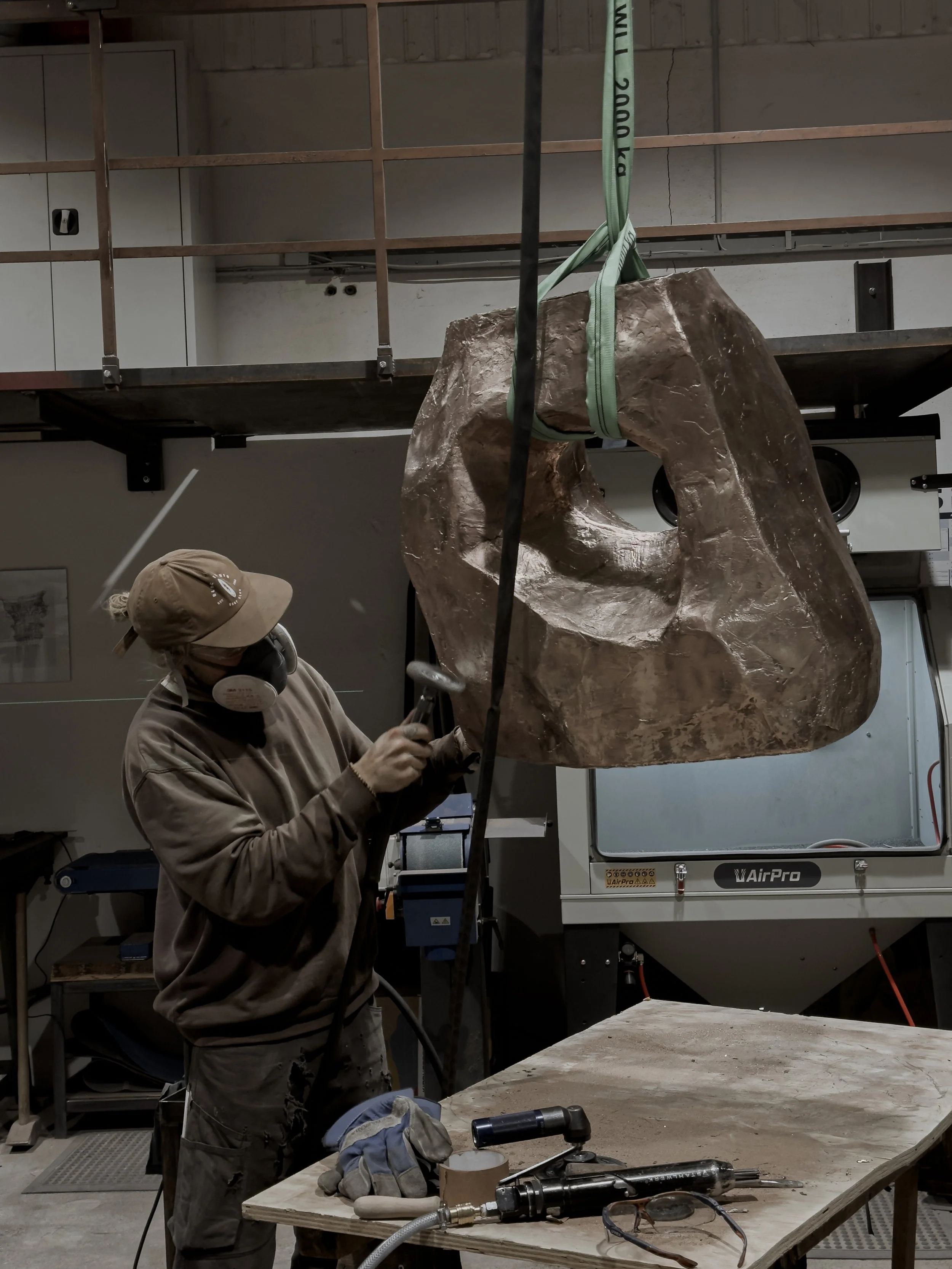 A person wearing a cap, face mask, and gloves is working with a grinder on a large, irregularly shaped metallic sculpture hanging from a strap in an art studio.