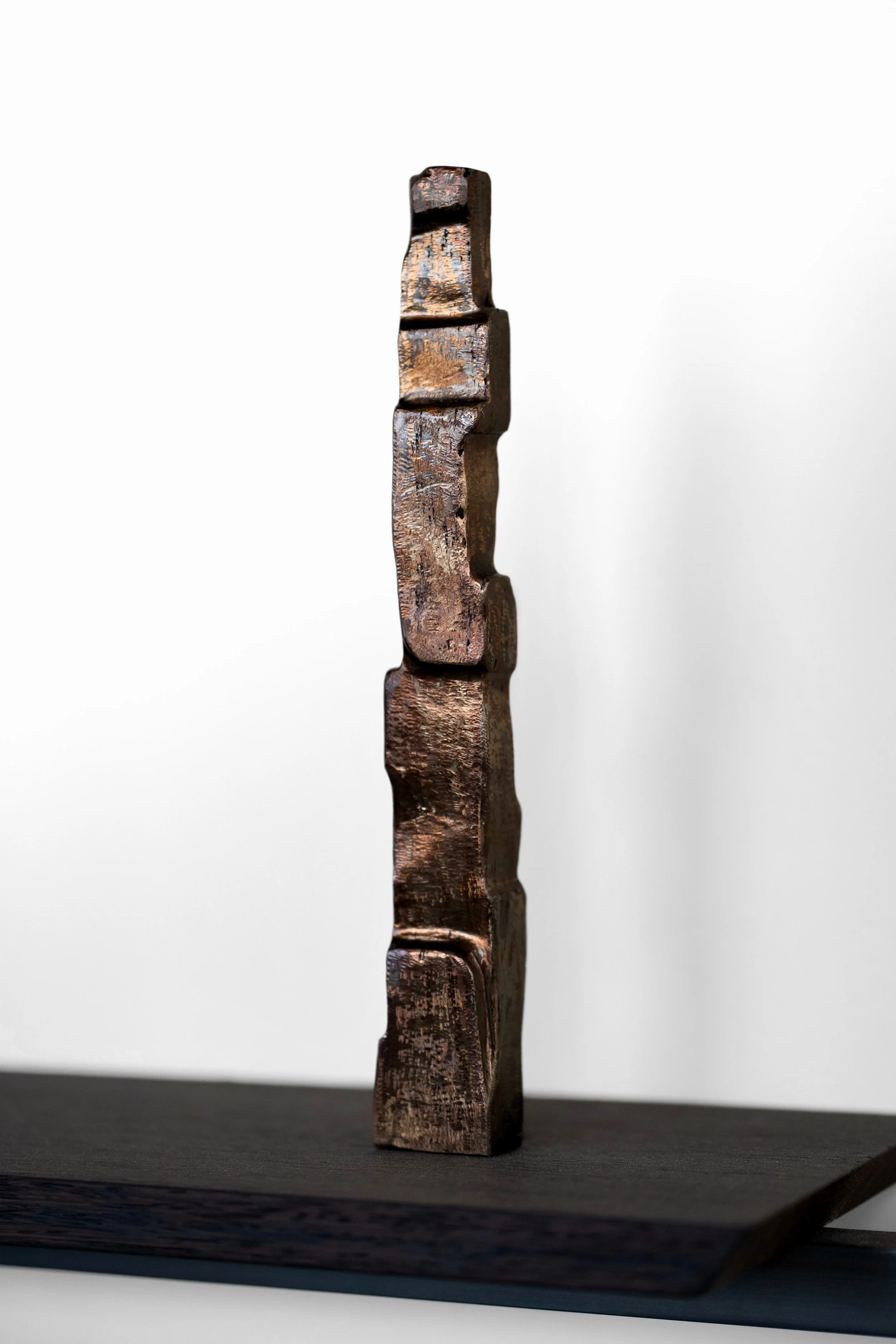 Vertical abstract bronze sculpture with a weathered dark brown finish, displayed on a black stand against a white background.