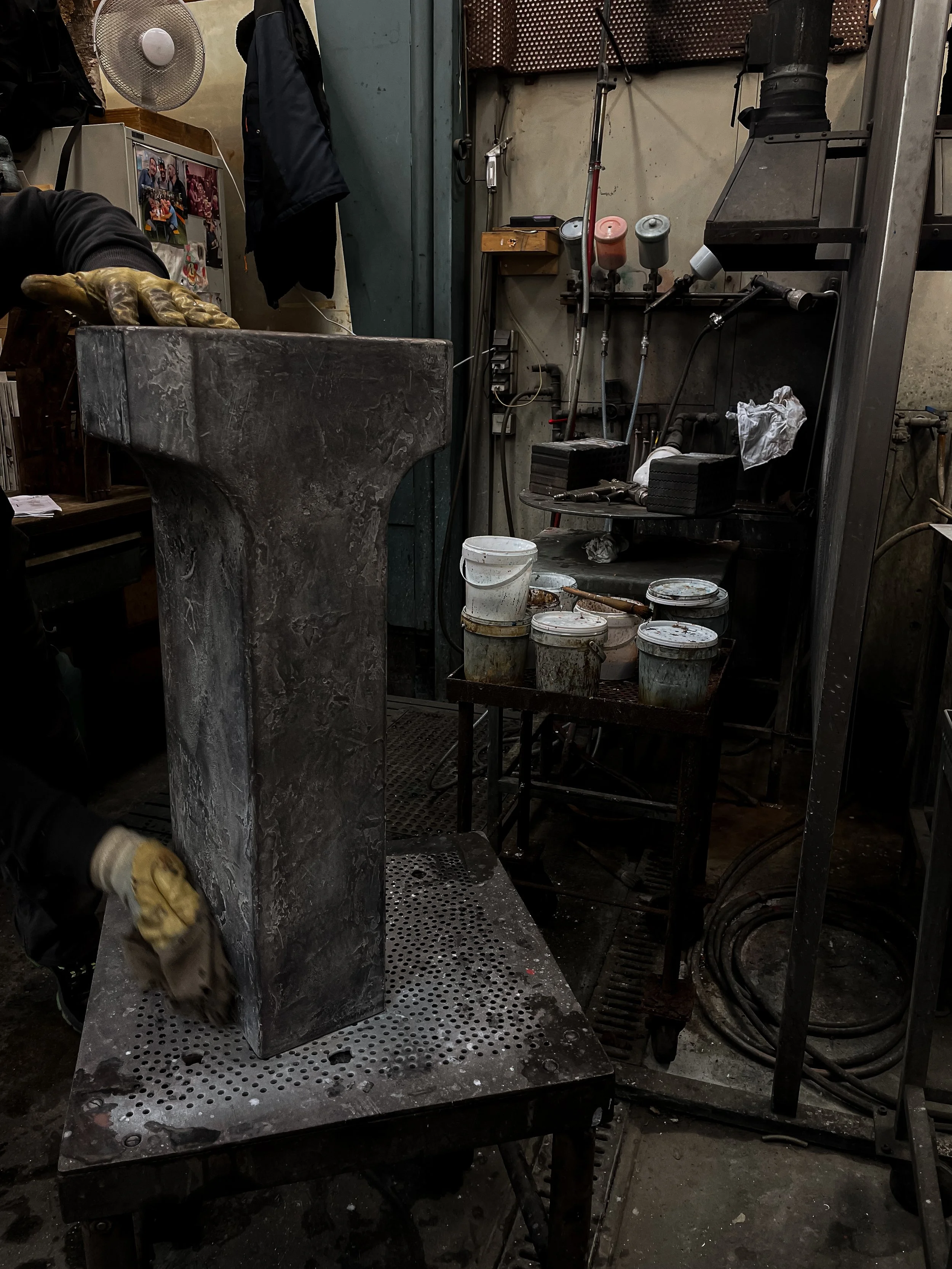 A worker wearing gloves handles a large, rectangular metal piece in a blacksmith workshop with various tools, buckets, and equipment visible around.