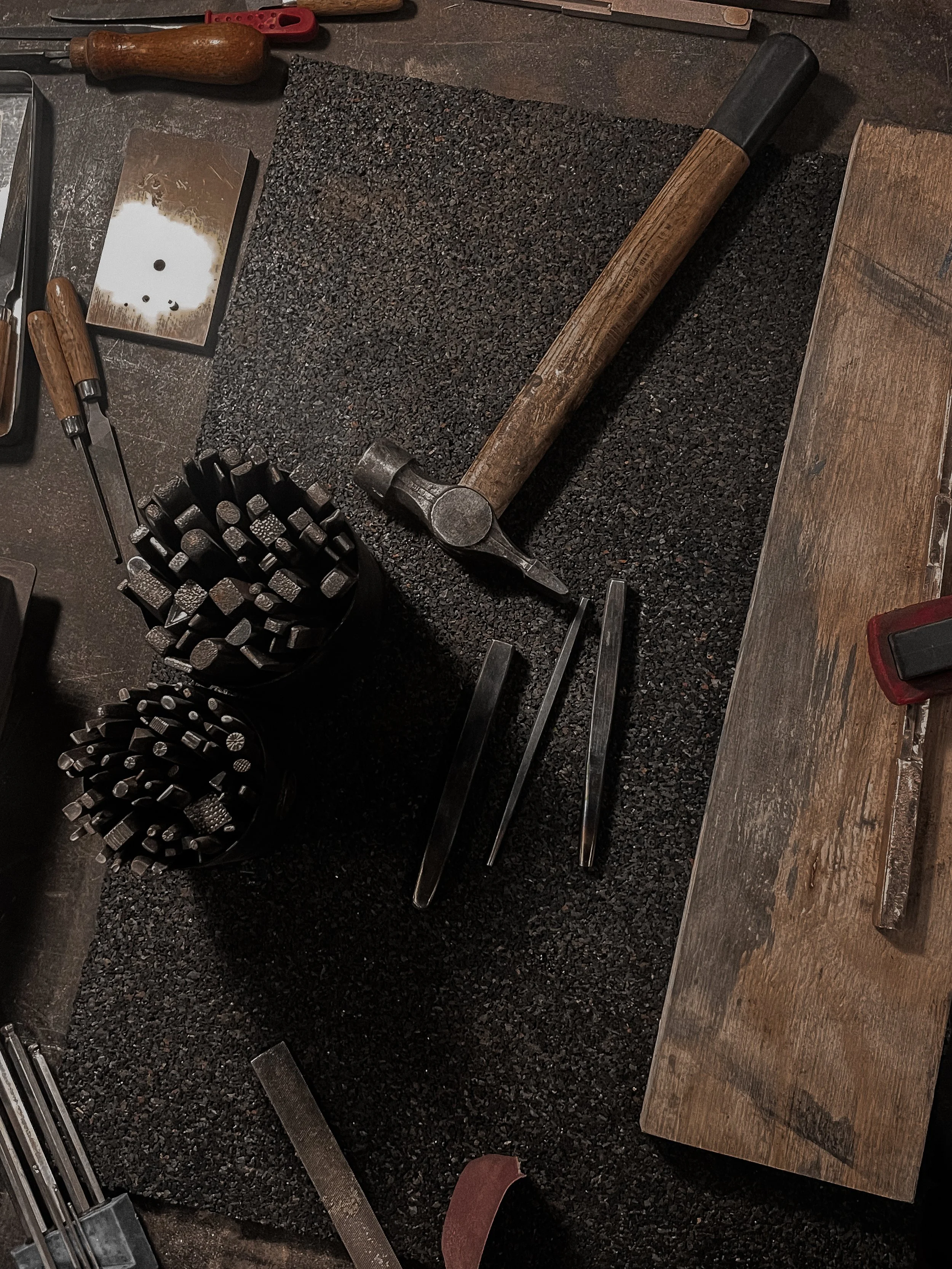 A workshop workspace with a hammer, tongs, chisels, and various hand tools on a black mat and wooden surface.
