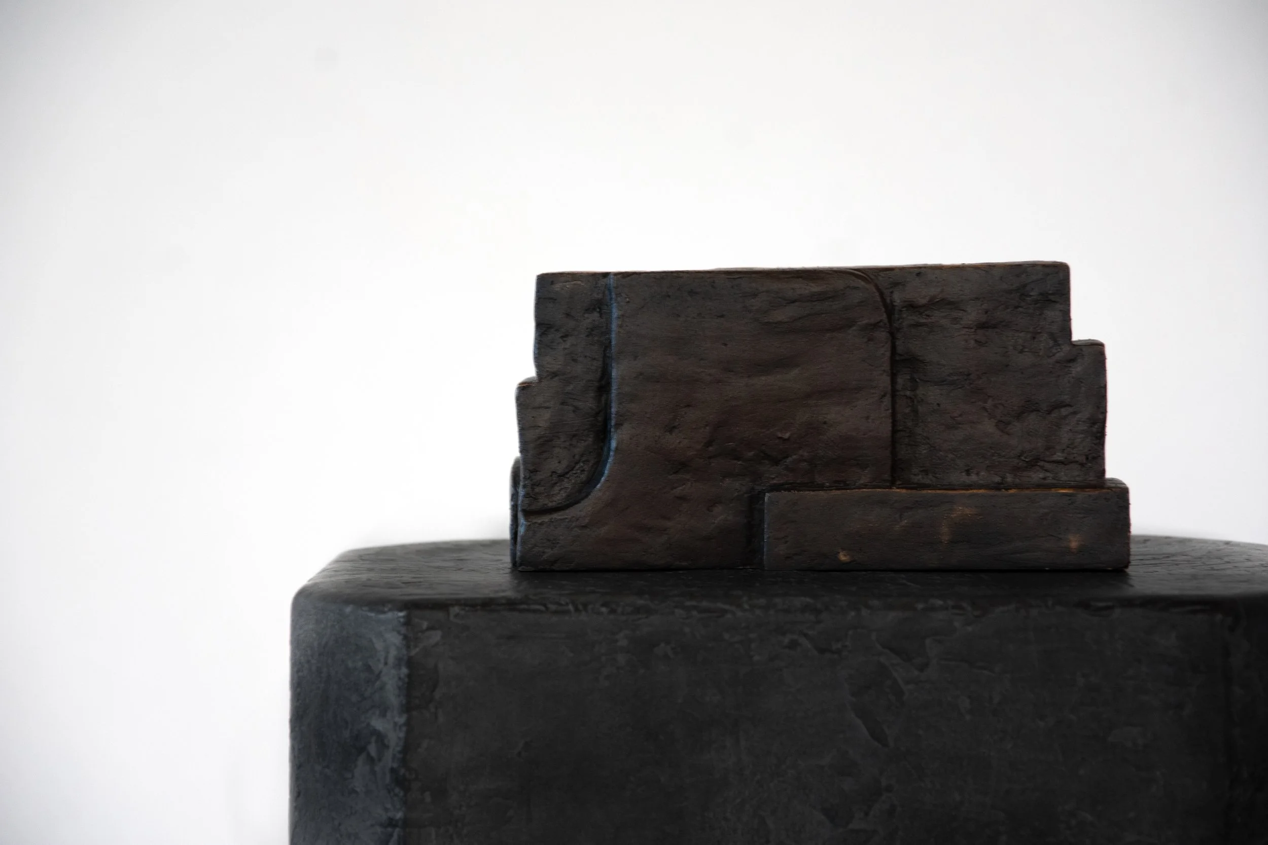 Dark textured rectangular sculpture on a dark pedestal against a plain white background.