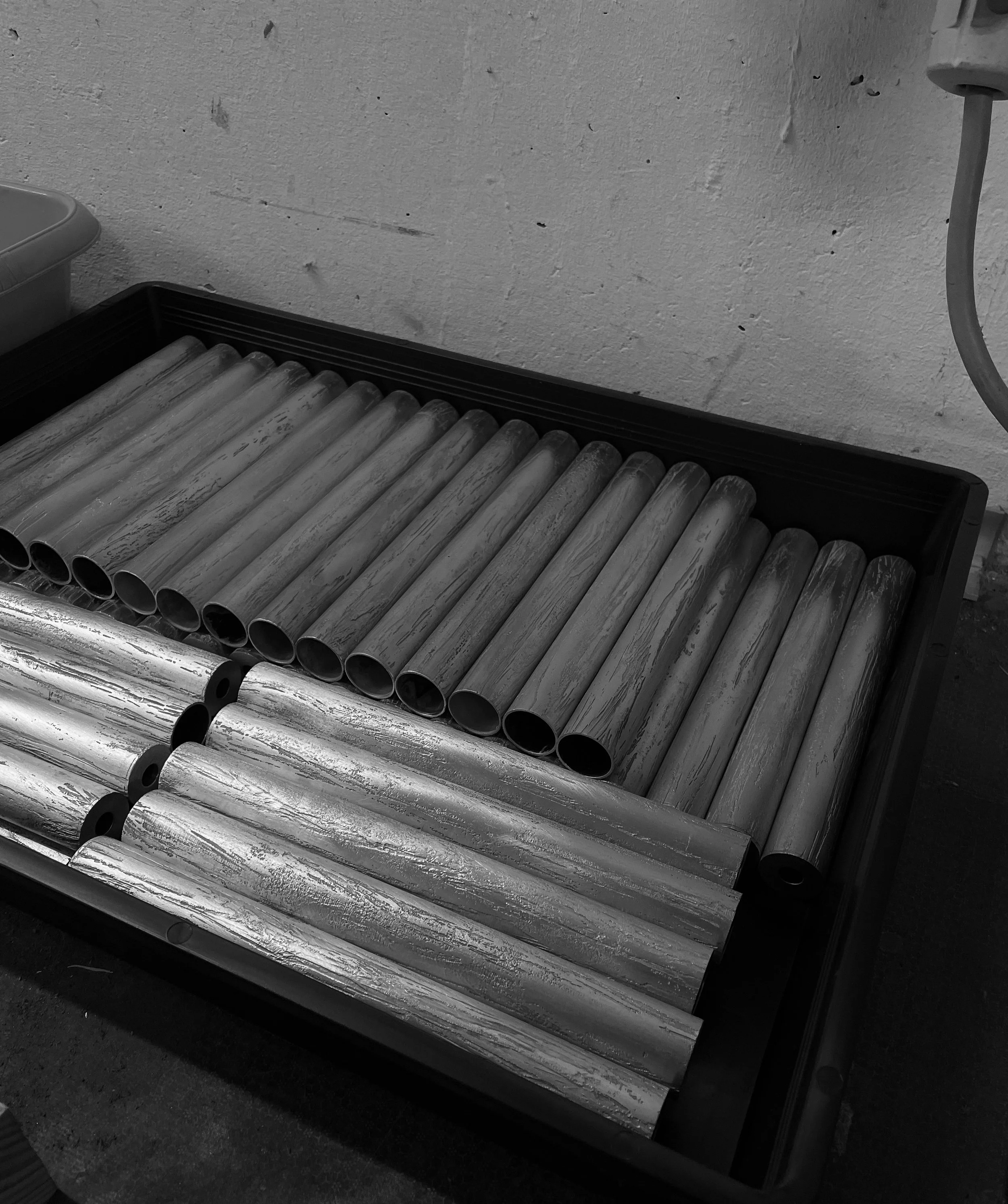 A tray of 23 metal pipes, 13 with a textured, weathered surface and 10 with a smooth, shiny surface, laid horizontally indoors against a textured wall.
