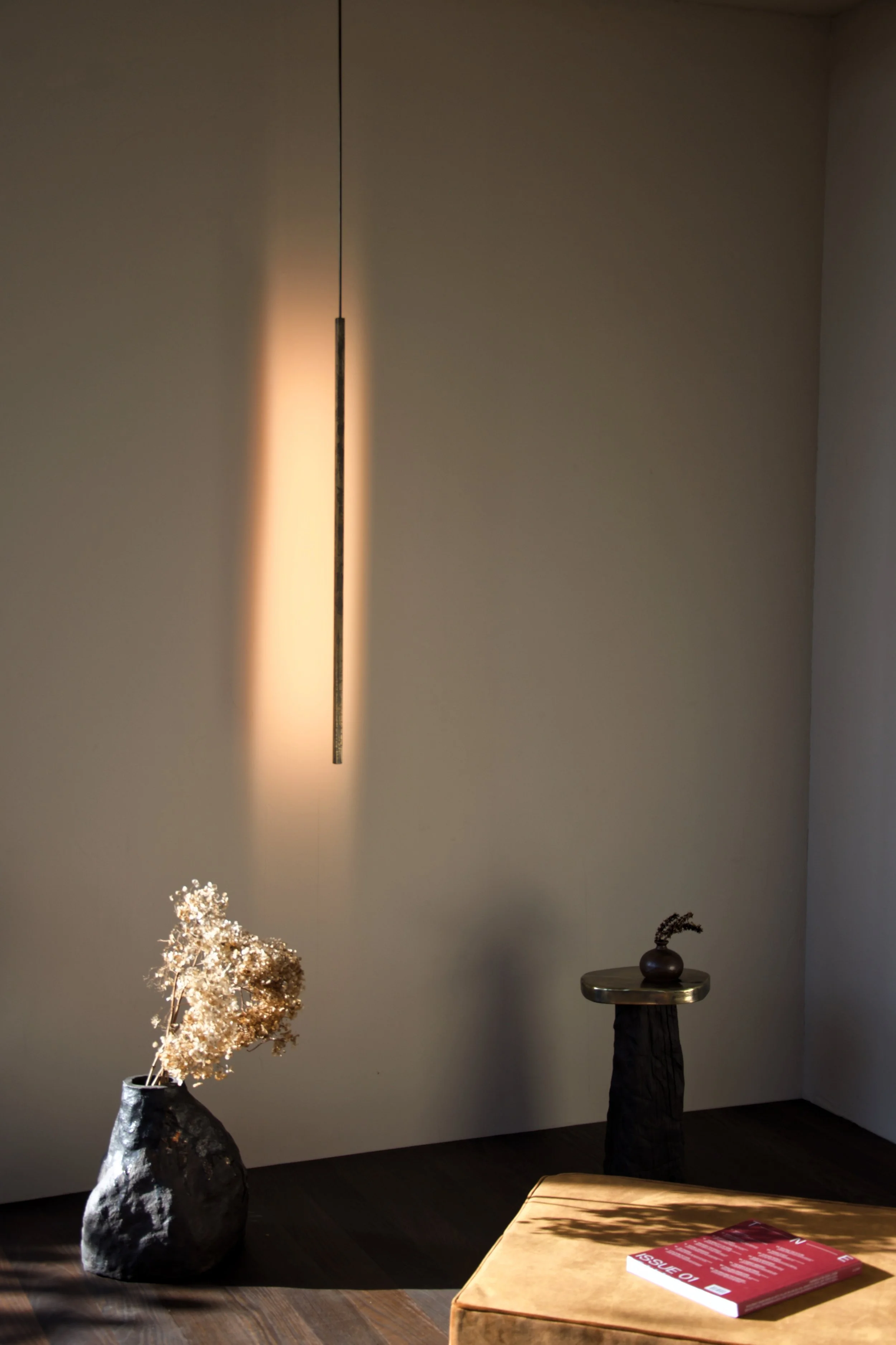 Minimalist interior with a hanging light fixture, a black vase with dried flowers, a small round table with a decorative vase, and a brown upholstered chair with a red magazine on top.