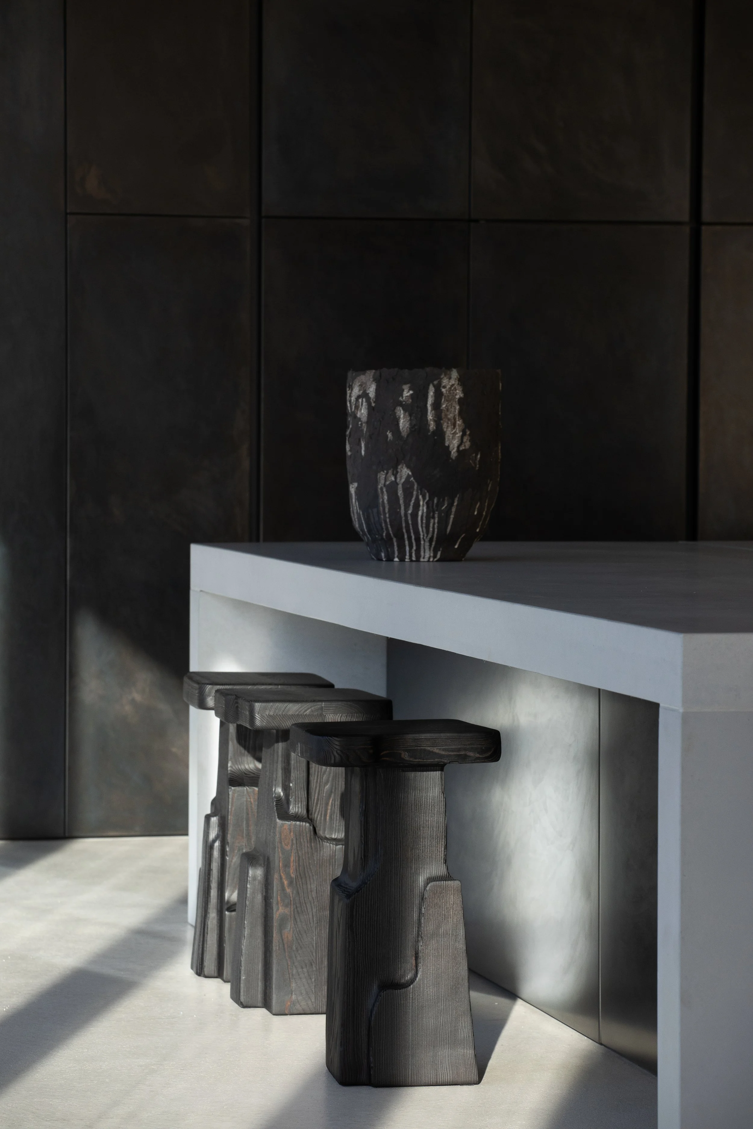 Modern interior with three black wooden stools under a white countertop, a dark decorative vase on the counter, and black framed wall panels.
