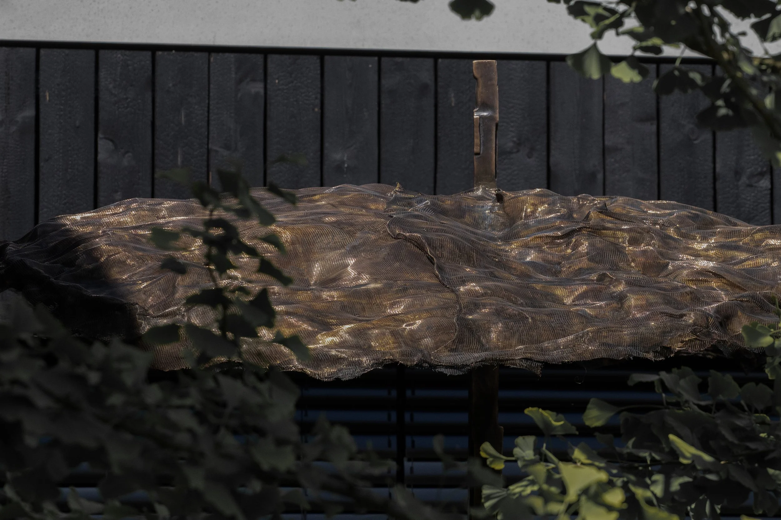 Outdoor patio with a large round table covered by a mesh umbrella, surrounded by greenery, with a black wooden fence in the background.