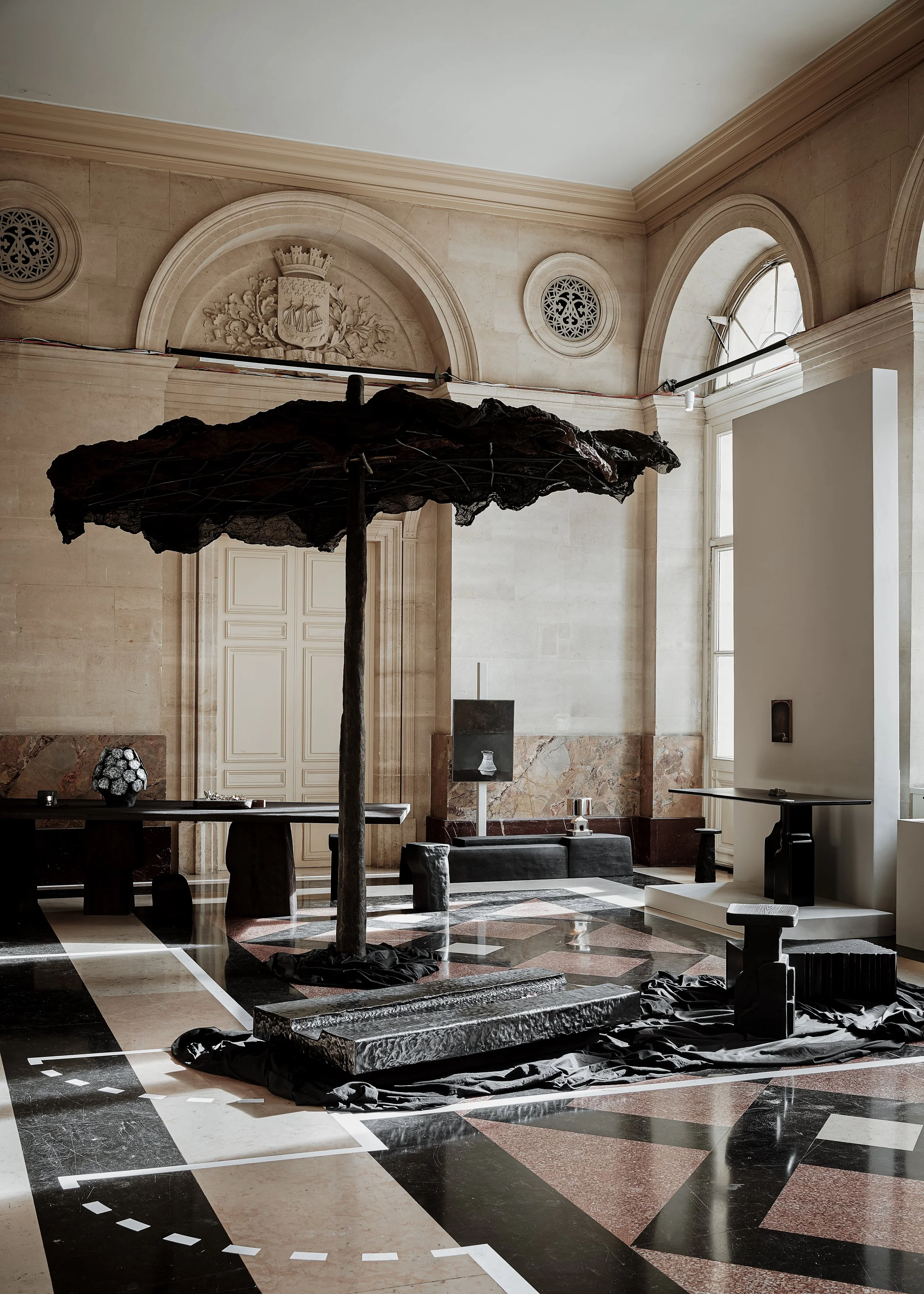 Interior of a historical building with classical architecture, high arched windows, and a decorative crest on the wall. Sculptural art installation featuring a large black umbrella-like structure with textured fabric and a central pole, surrounded by black fabric and various black objects on the floor.