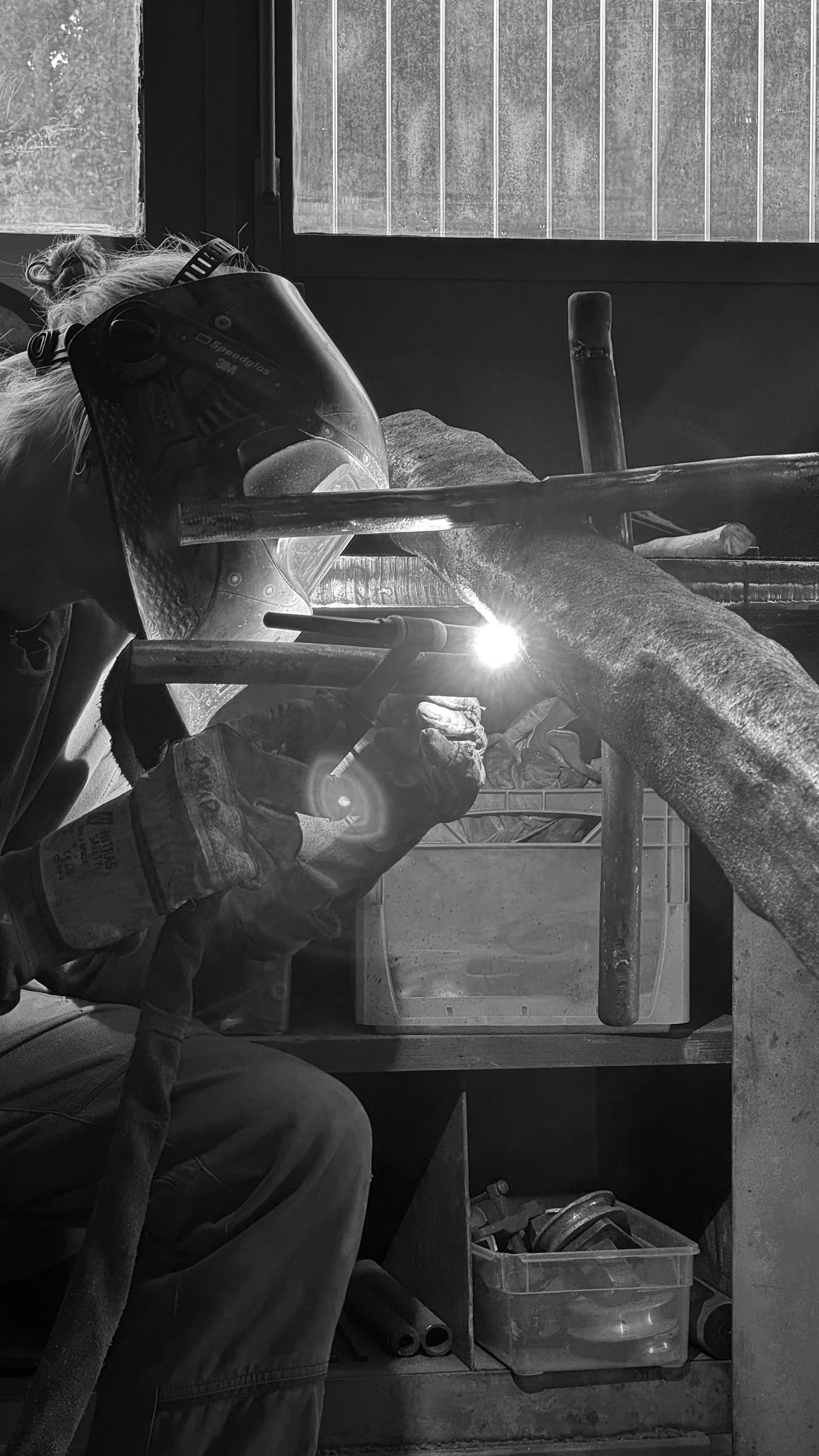 A person welding metal, wearing protective gloves and a welding helmet, in a workshop with tools and equipment around.