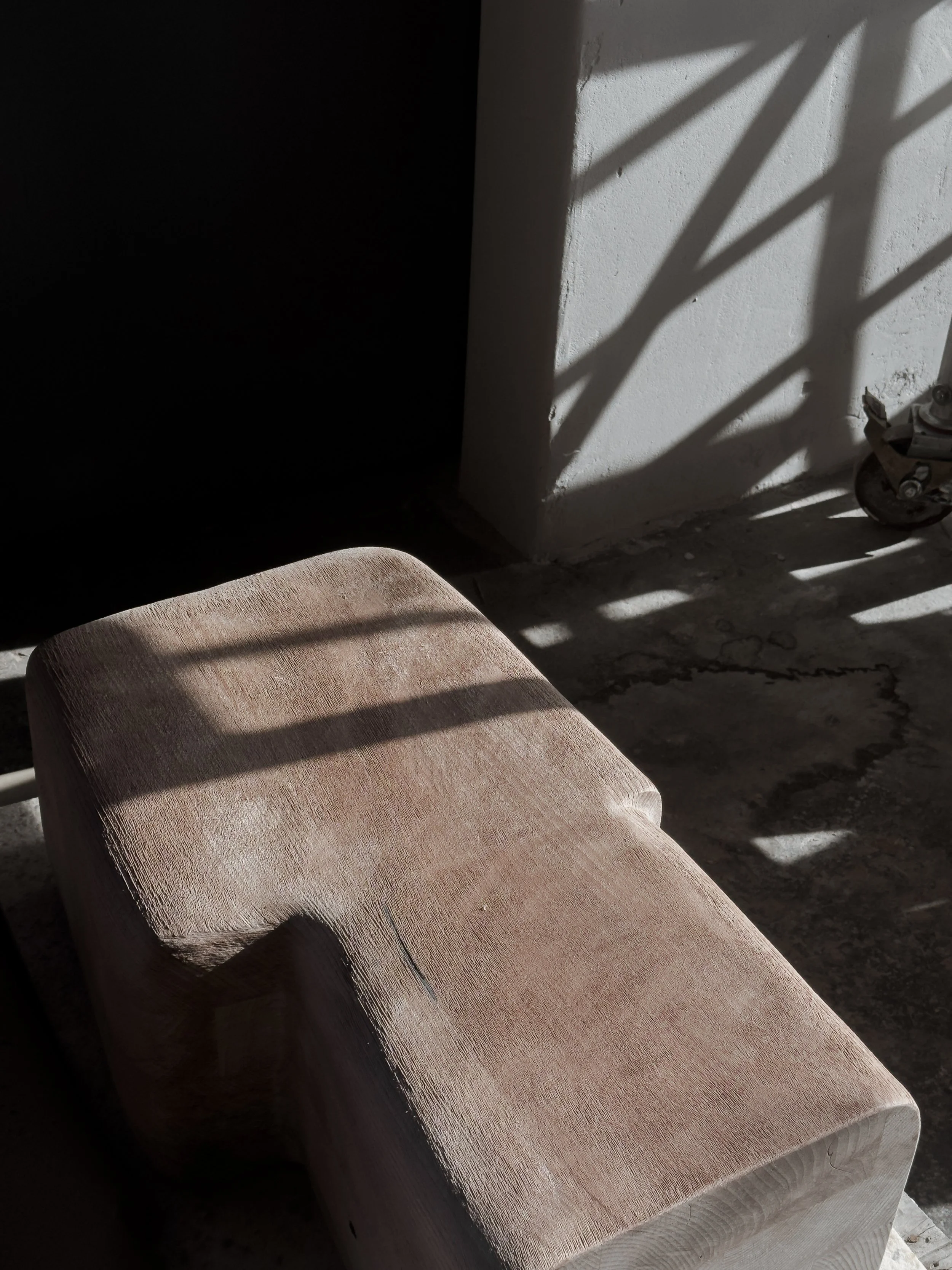 A wooden stool or bench in sunlight casting shadows on the wall and floor, with a roller skate partially visible in the background.