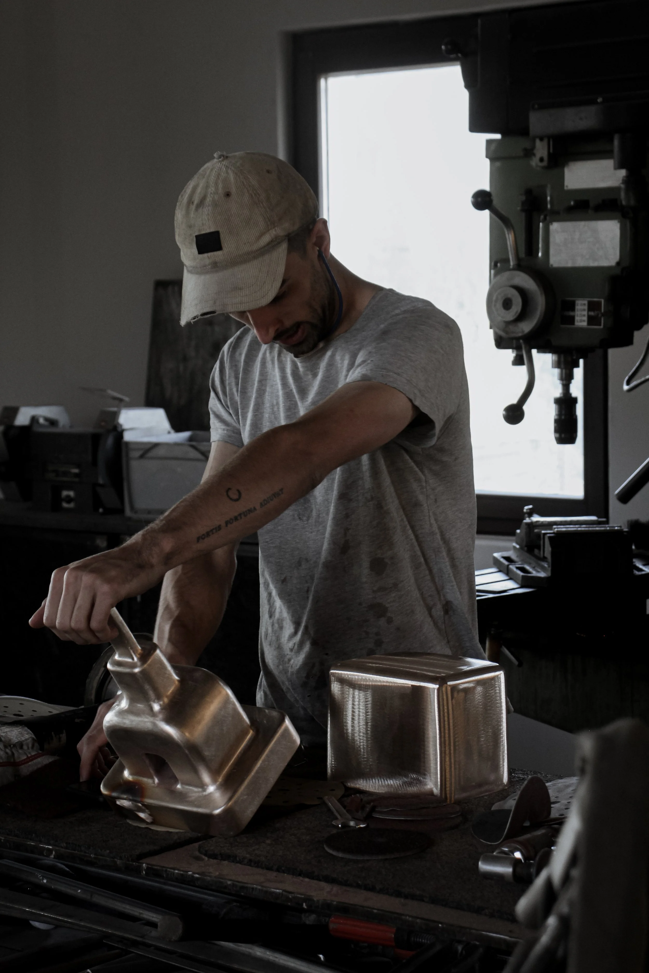 A man in a grey t-shirt and beige cap working in a workshop, handling metal molds or blocks, with machinery and a window in the background.