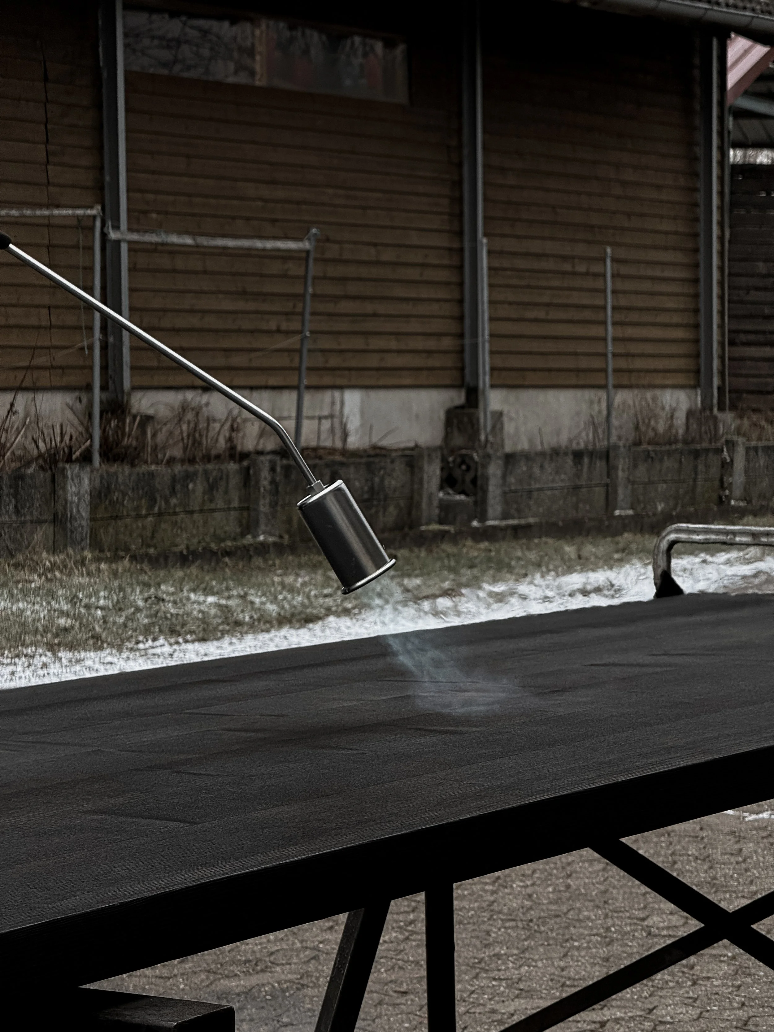 A small handheld fogging machine emitting mist onto a black surface outdoors with a wooden fence and grass in the background.