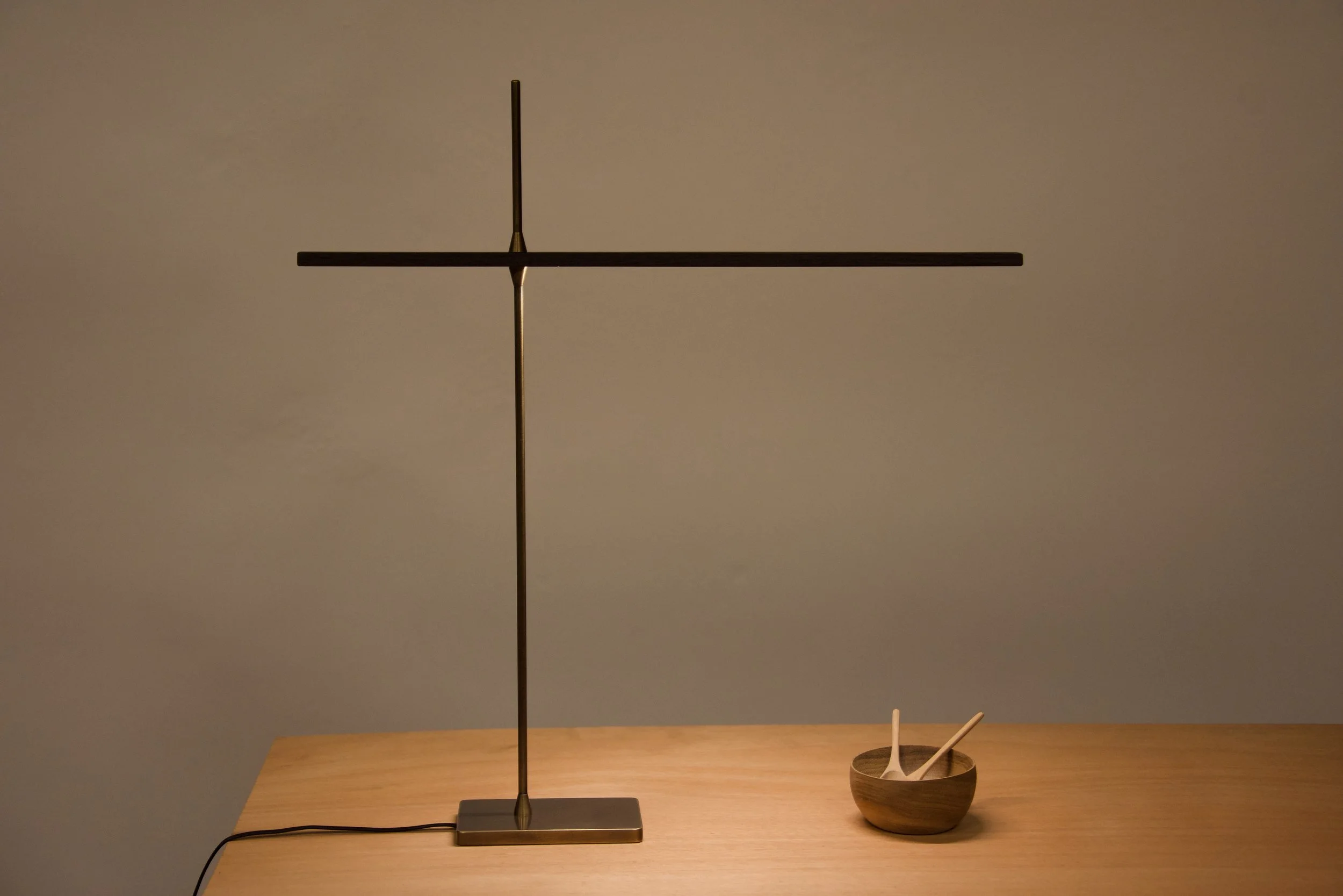 A minimalist table lamp with a thin metal stand and horizontal arm, turned off, on a wooden surface next to a small wooden bowl holding two wooden spoons.