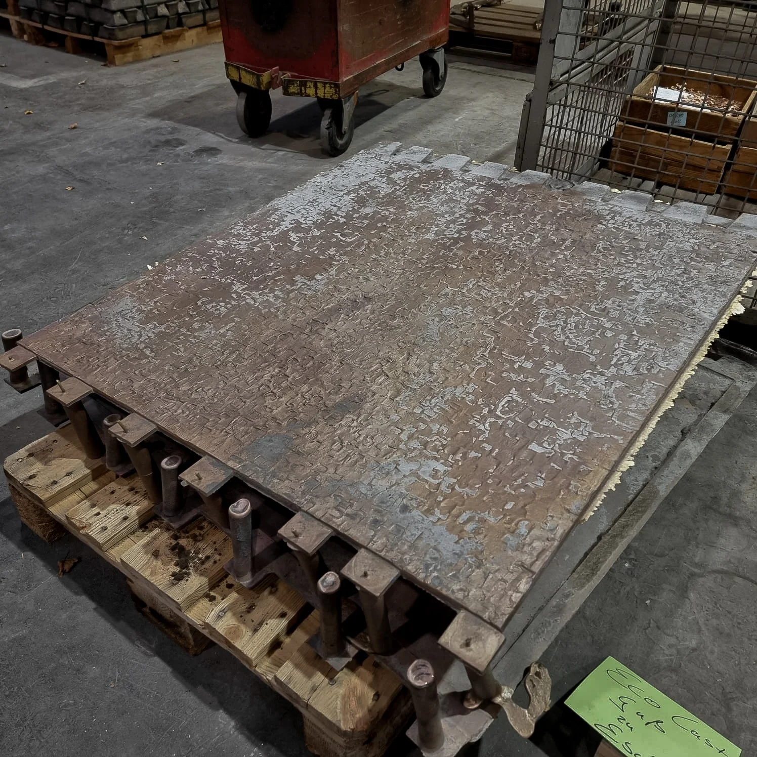 A large, rusted, metal industrial mold rests on a wooden pallet in a workshop, surrounded by tools and equipment.