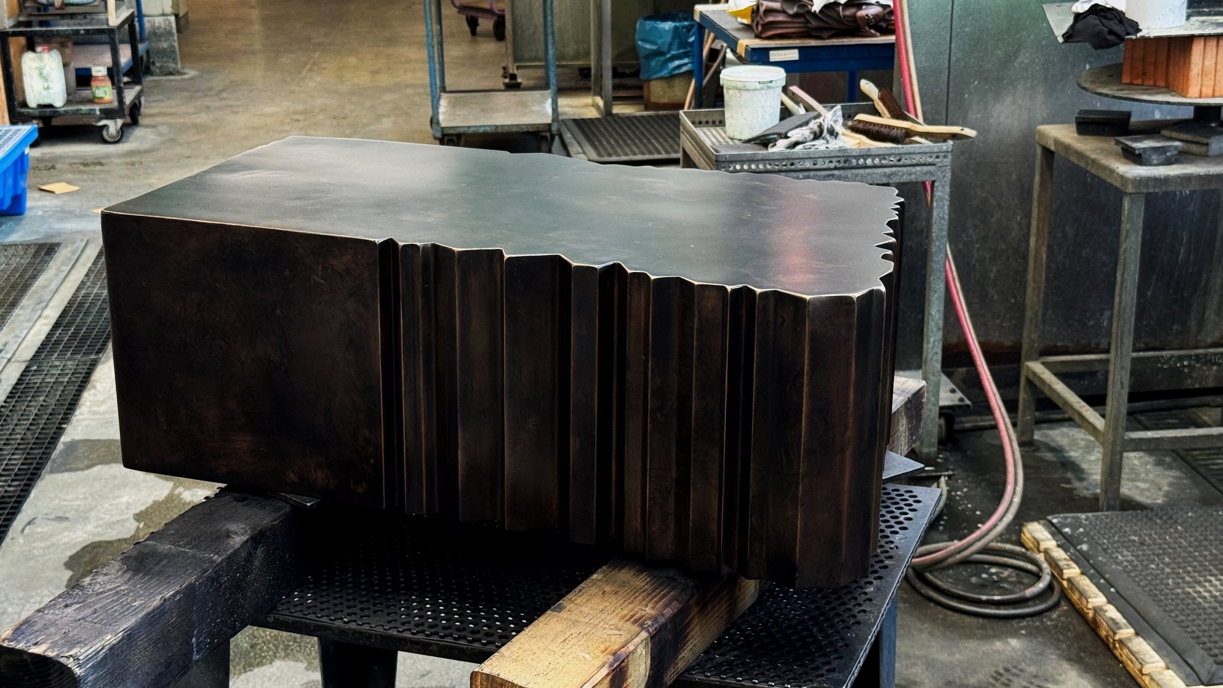A large, dark-colored, carved piece of metal, polished top and decorative vertical grooves along the side rests on a workbench in a workshop.