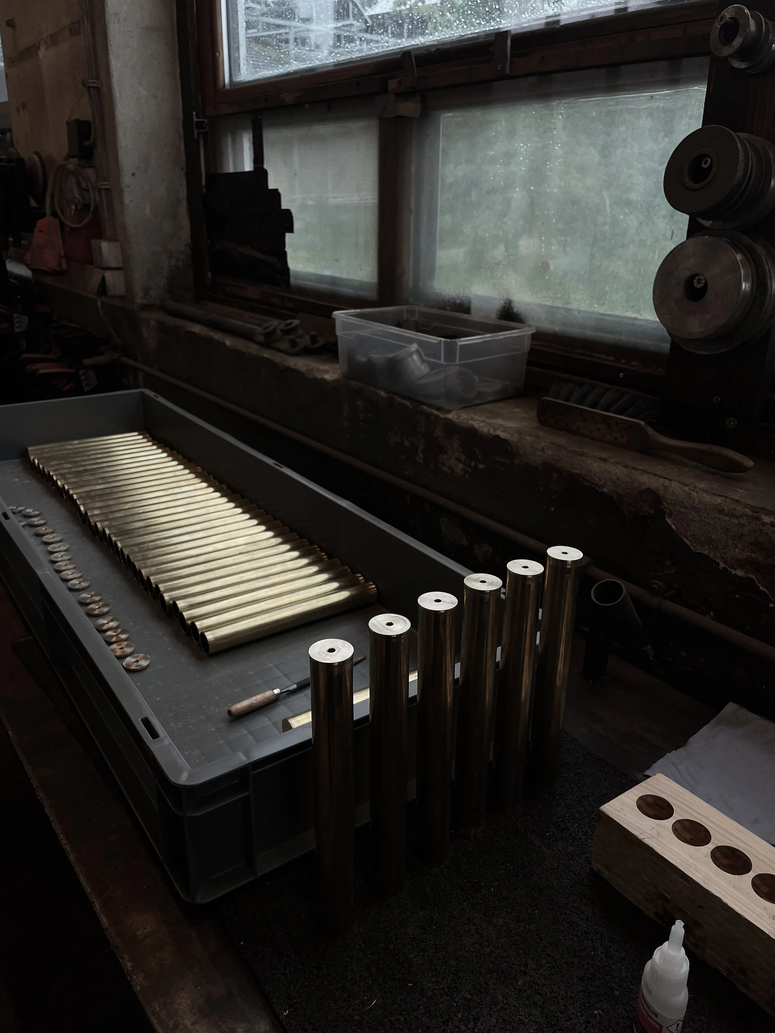 Workshop with brass tubes and metal components on a workbench, surrounded by tools and supplies, with a window showing an outdoor scene.