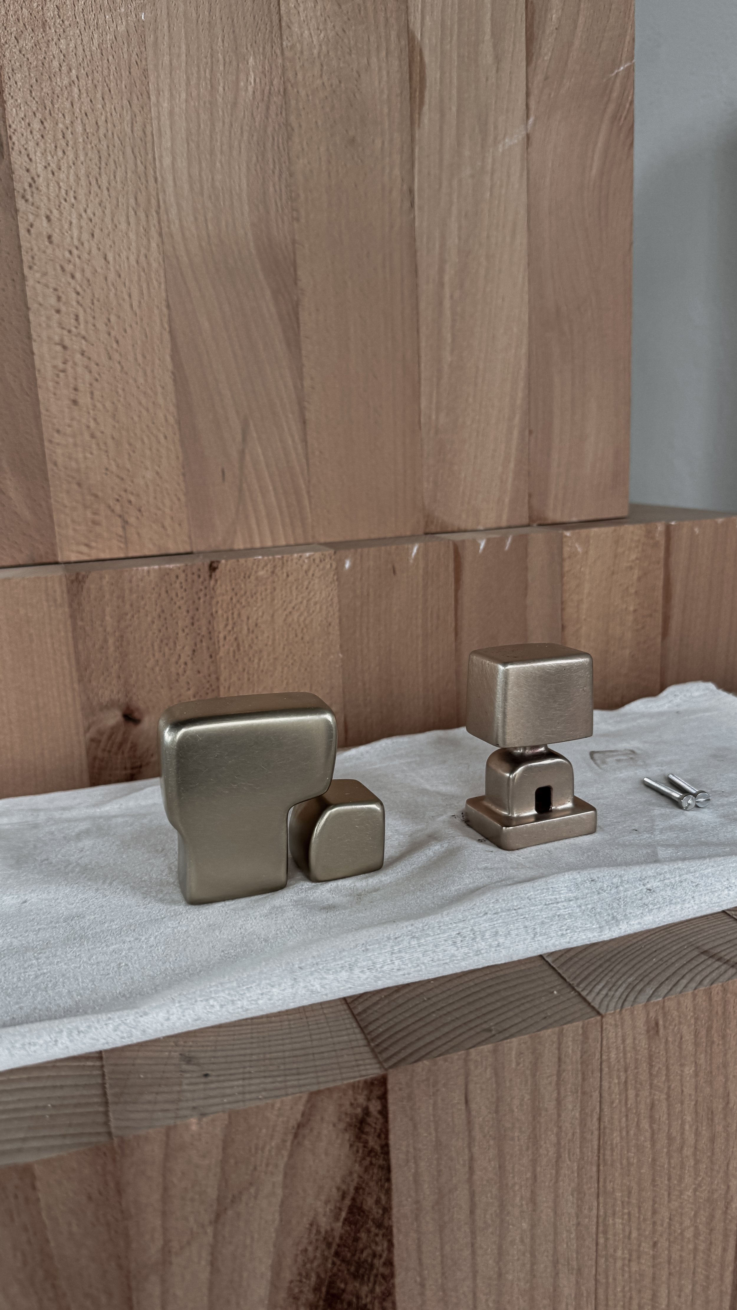 Two metallic cabinet knobs, one larger and one smaller, placed on a white cloth, with two screws lying next to them on a wooden surface