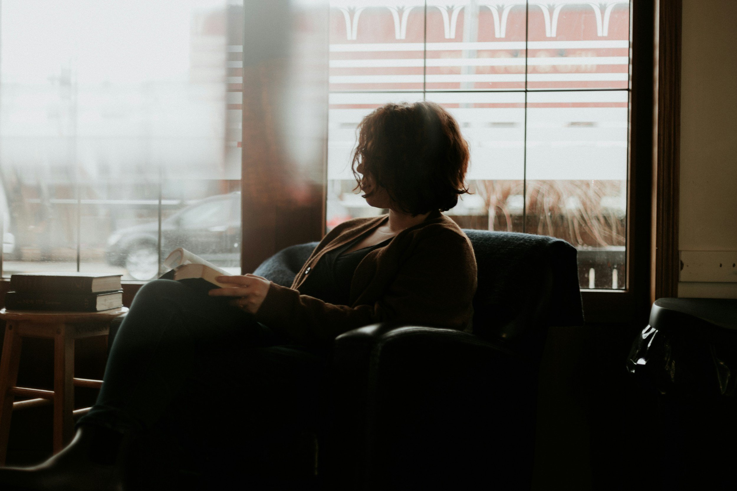 A woman with curly hair is sitting inside a coffee shop, reading a book near a window.