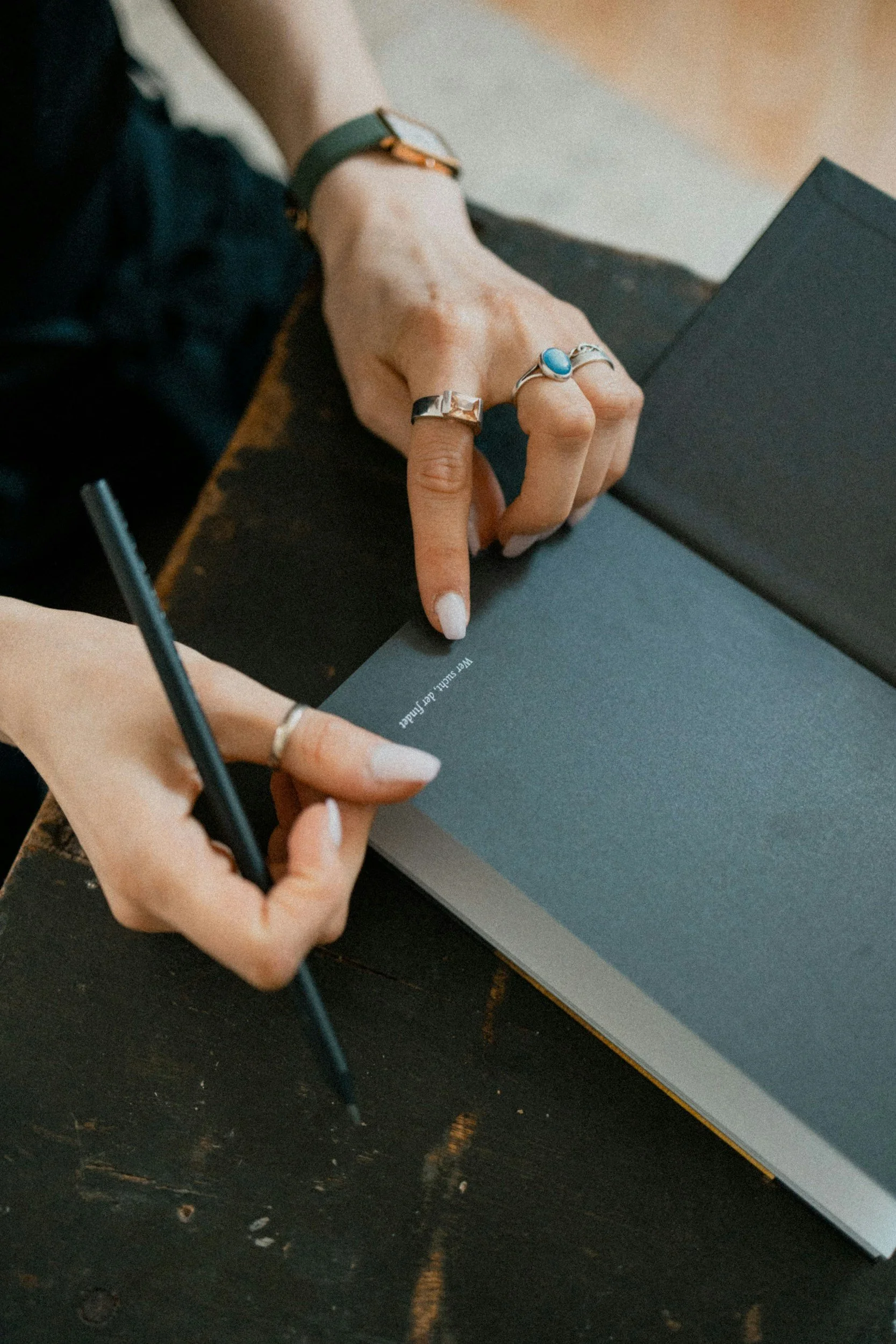 Close-up of a person's hand touching a black book or folder with a silver ring on their middle finger, a blue ring on their index finger, a black wristband, and a gold bracelet. The person is sitting at a wooden table with a dark wood surface, and a pen is visible nearby.