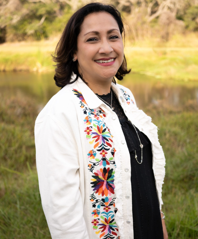 Woman outdoors with a smile, wearing a white embroidered jacket and black top, near a lake and trees.