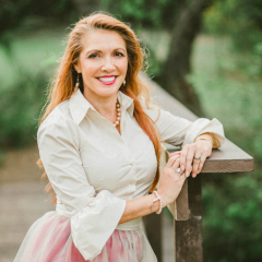 A smiling woman with long red hair in a white blouse and pink skirt, standing outdoors next to a wooden railing with a blurred green background.